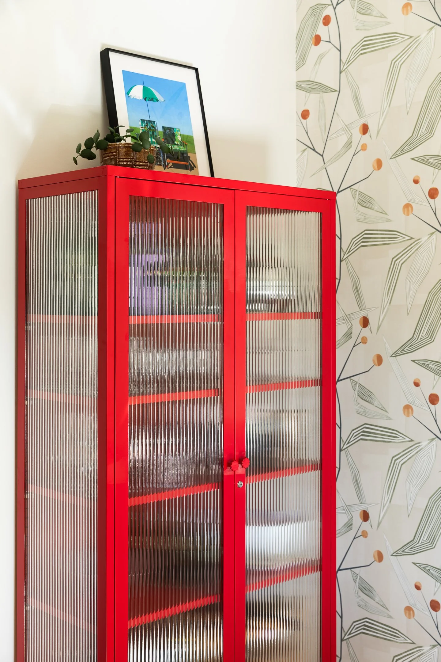 A red cabinet with ridged glass doors, topped with a framed art print and a small plant inside a basket.