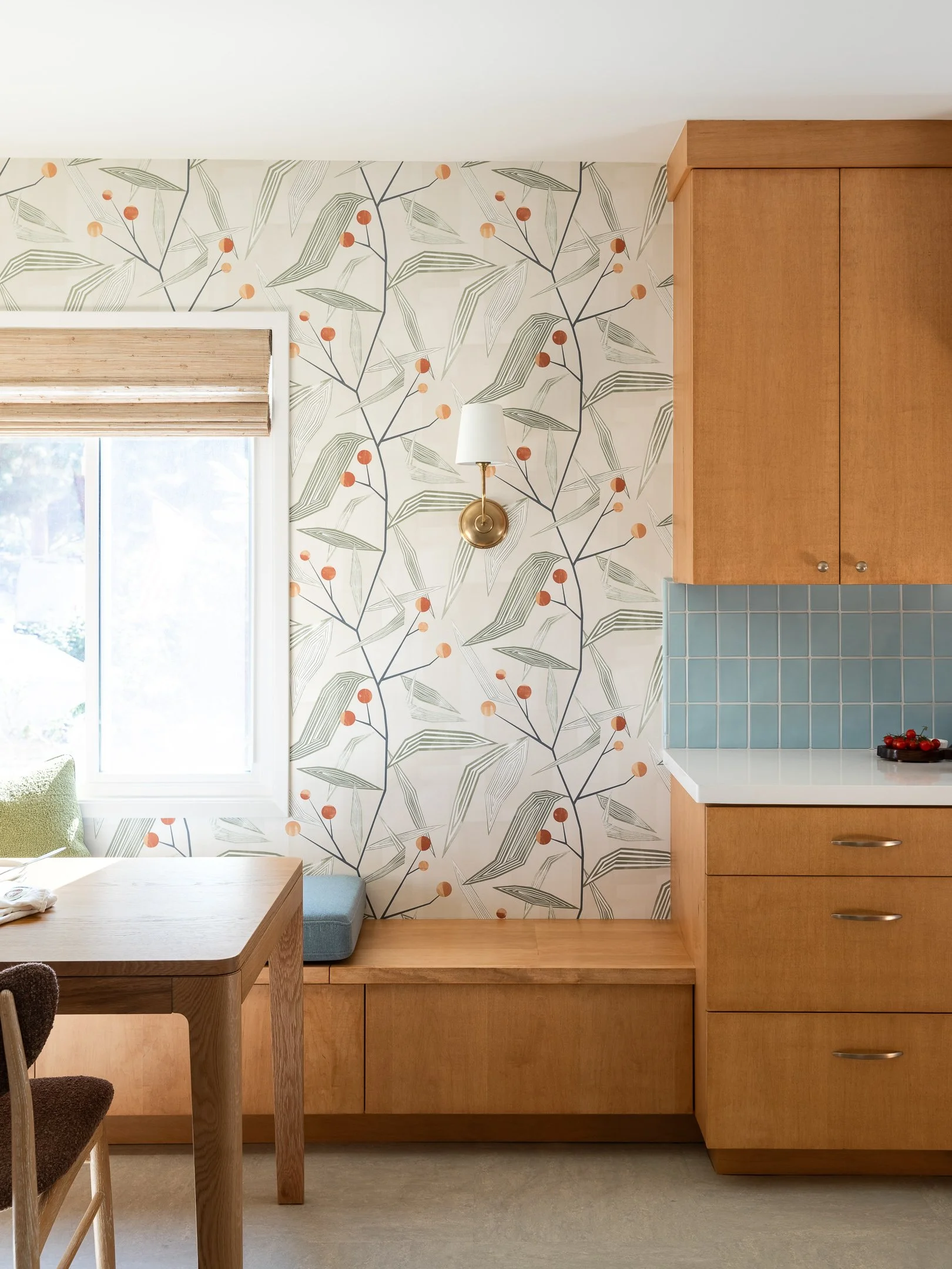 Interior view of a kitchen or dining area showcasing a wooden bench, table, window with a bamboo shade, wall with botanical wallpaper, a brass wall sconce with a white shade, upper wooden cabinets, and a countertop with a bowl of fruit.