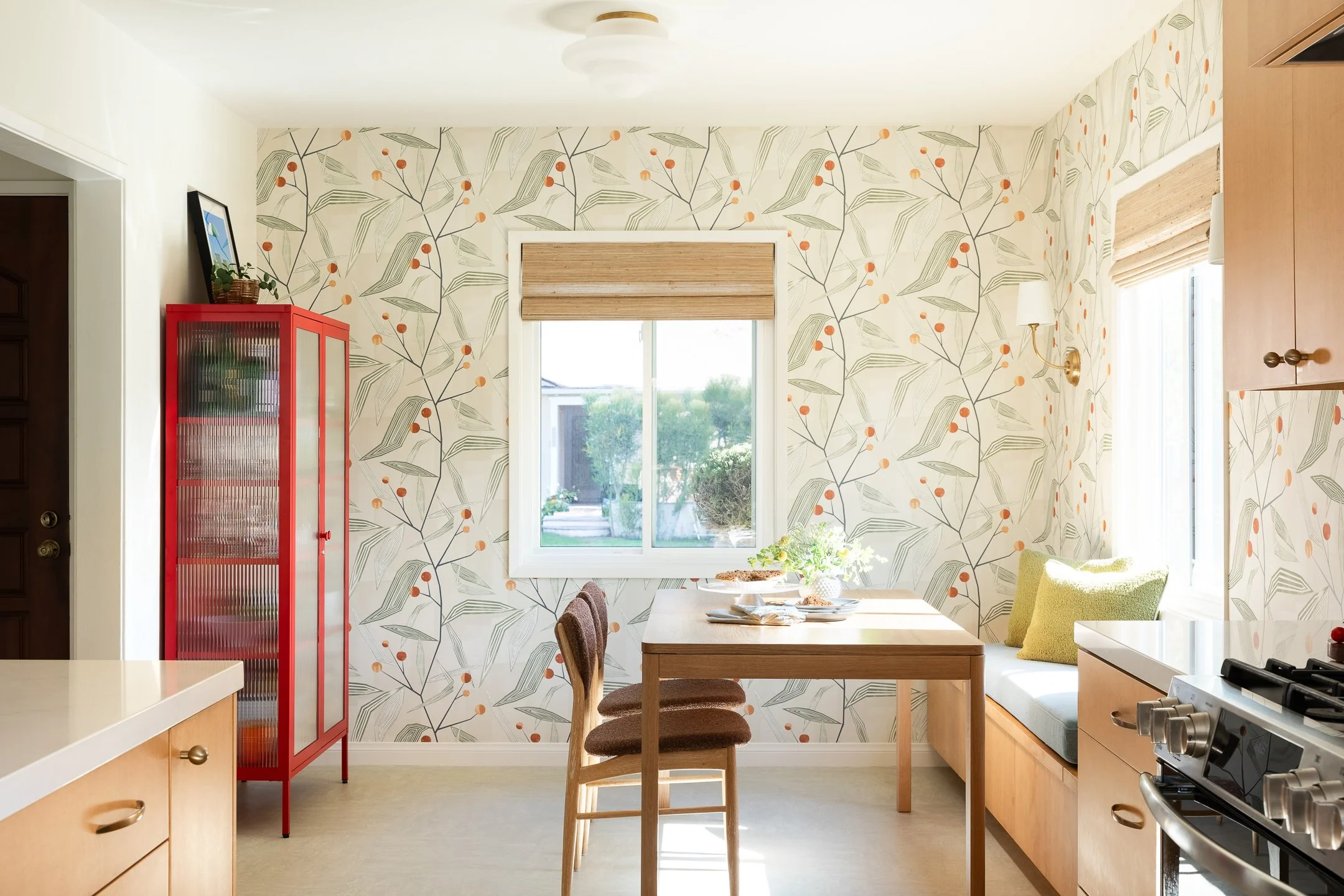 Bright kitchen with wallpaper featuring leaf and berry pattern, two windows letting in natural light, a red cabinet, a wooden dining table with chairs, and a window bench with pillows.