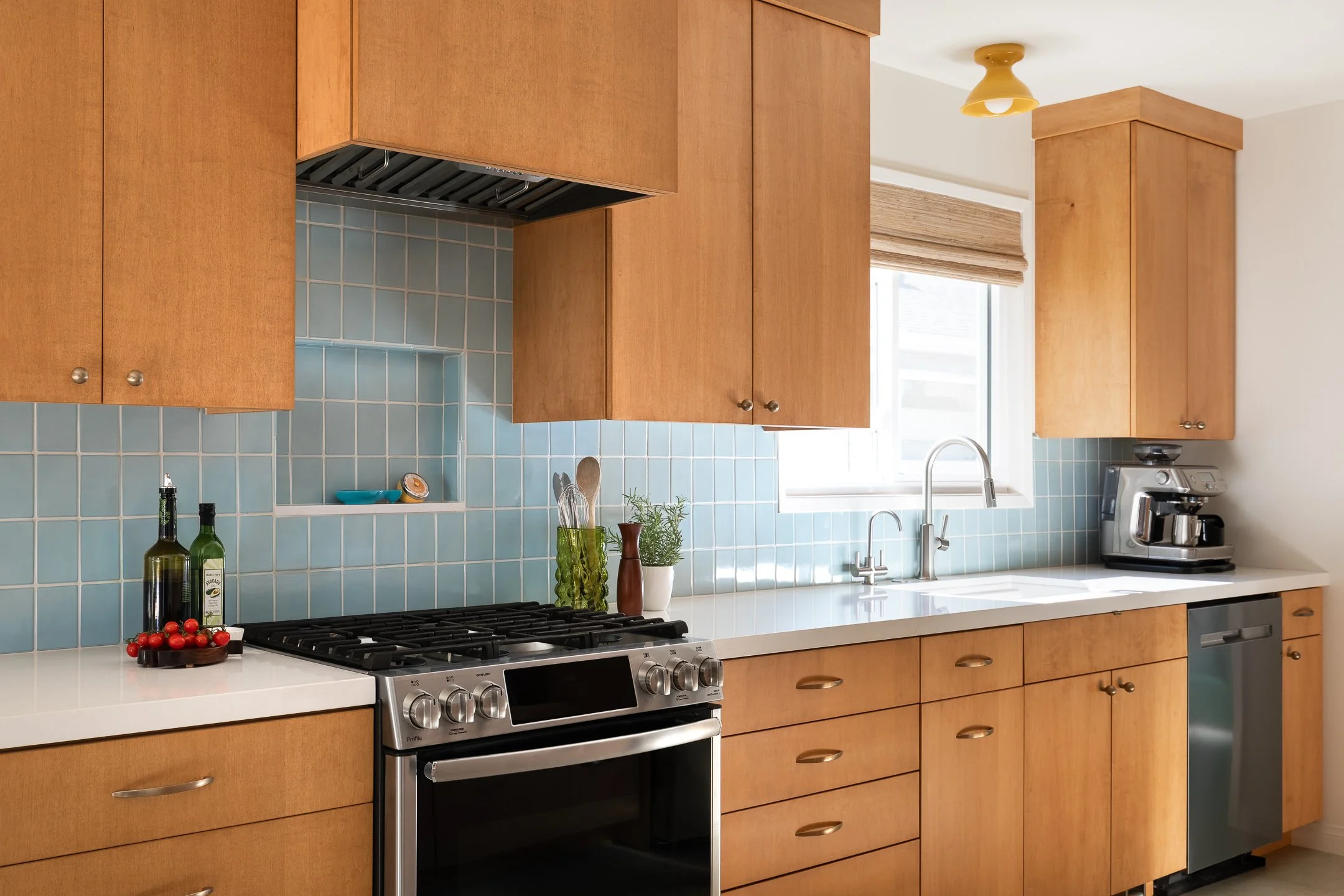 Kitchen with wooden cabinets, light blue tiled backsplash, white countertop, gas stove, sink, and coffee maker, sunlight coming through a window.