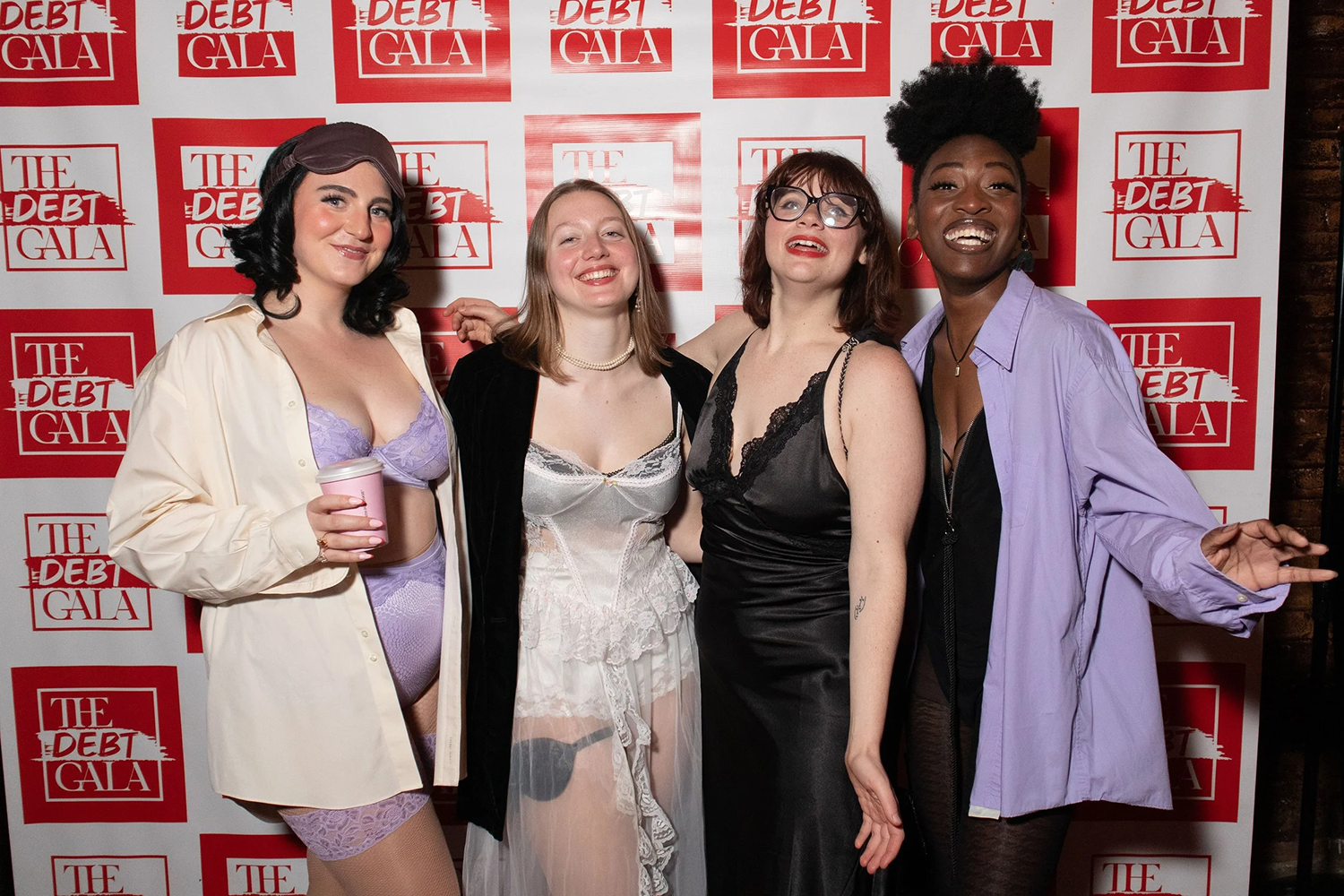Four people smiling having fun, to the left in purple lingerie, next to the right wearing white lingerie, next to the right wearing black lingerie and far right wearing black undergarments with purple overshirt