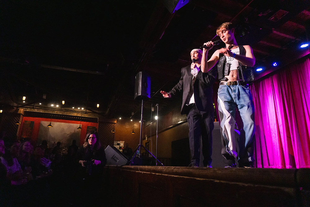 Presenting two males on stage performing by singing and dancing, one male with microphone in hand in jeans, leather vest and white crop top, other man in black dress pants and dress jacket with white undershirt dancing