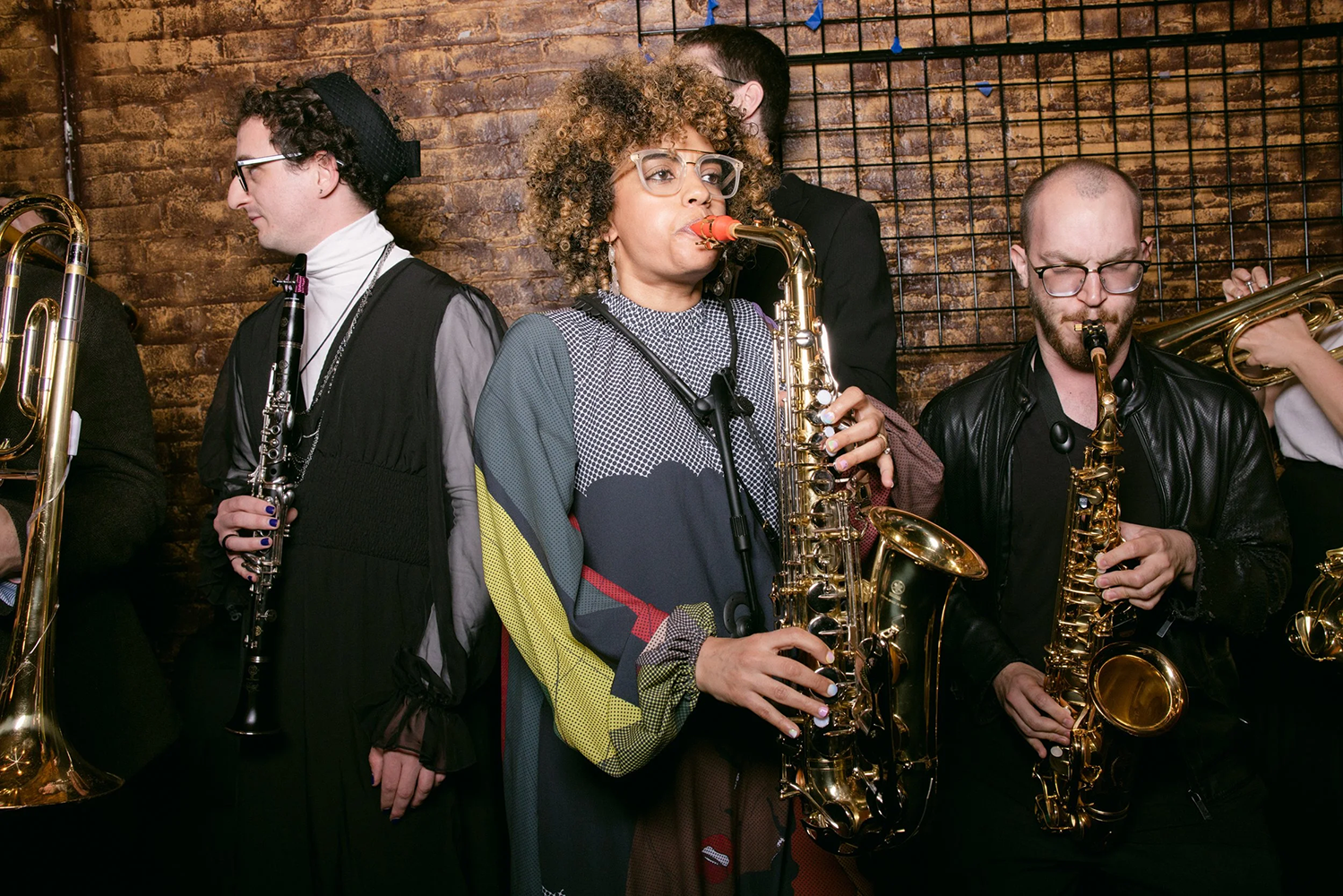 Group of people playing instruments such as flute, saxophone and trumpet, main focus on presenting woman playing saxophone