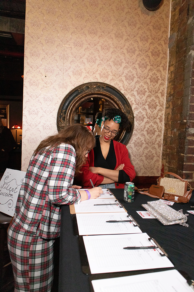 One person in red sweater, black shirt and turquoise-green bandana monitoring signup sheets for The Debt Gala and other person signing up with plaid suit and long hair