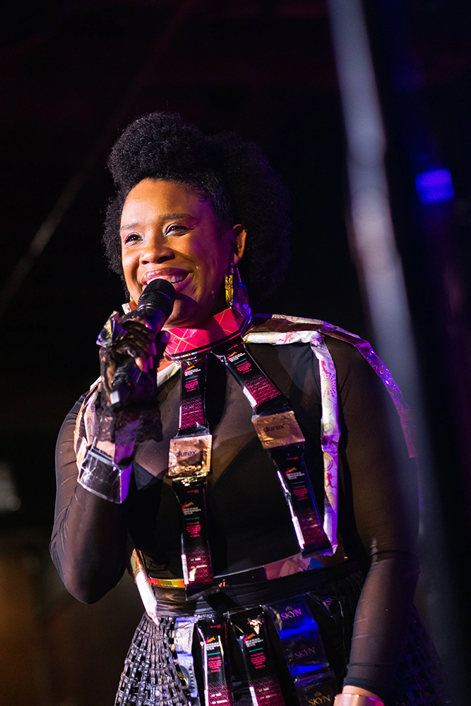 Presenting woman with condoms and tampons on outfit with black, shear underneath holding microphone performing and smiling