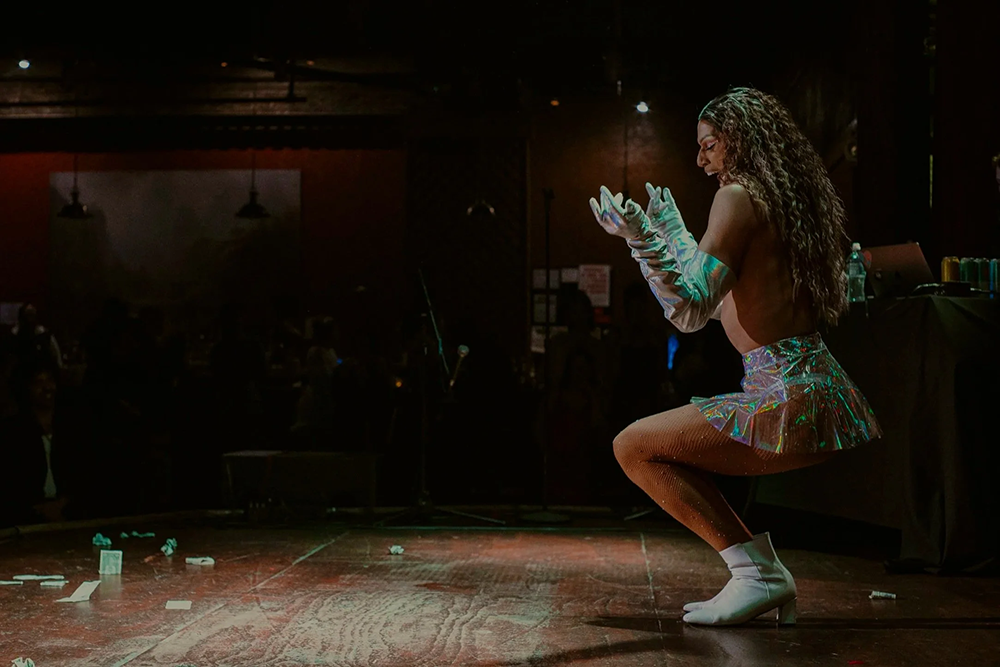 Person duck walking with fishnet, glittery stockings, white shoes with short heels, iridescent clear skirt and white, iridescent long gloves, long hair and smiling having fun