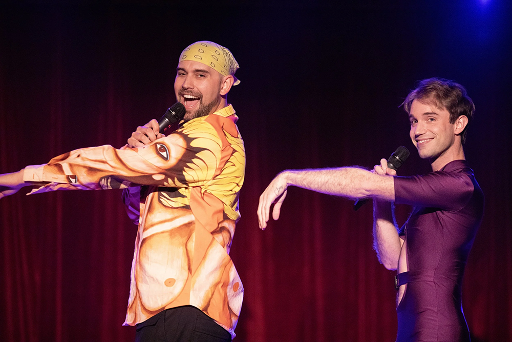 Two presenting men performing on stage, one man in looser fitting button down shirt and black pants, other in form fitting purple leotard