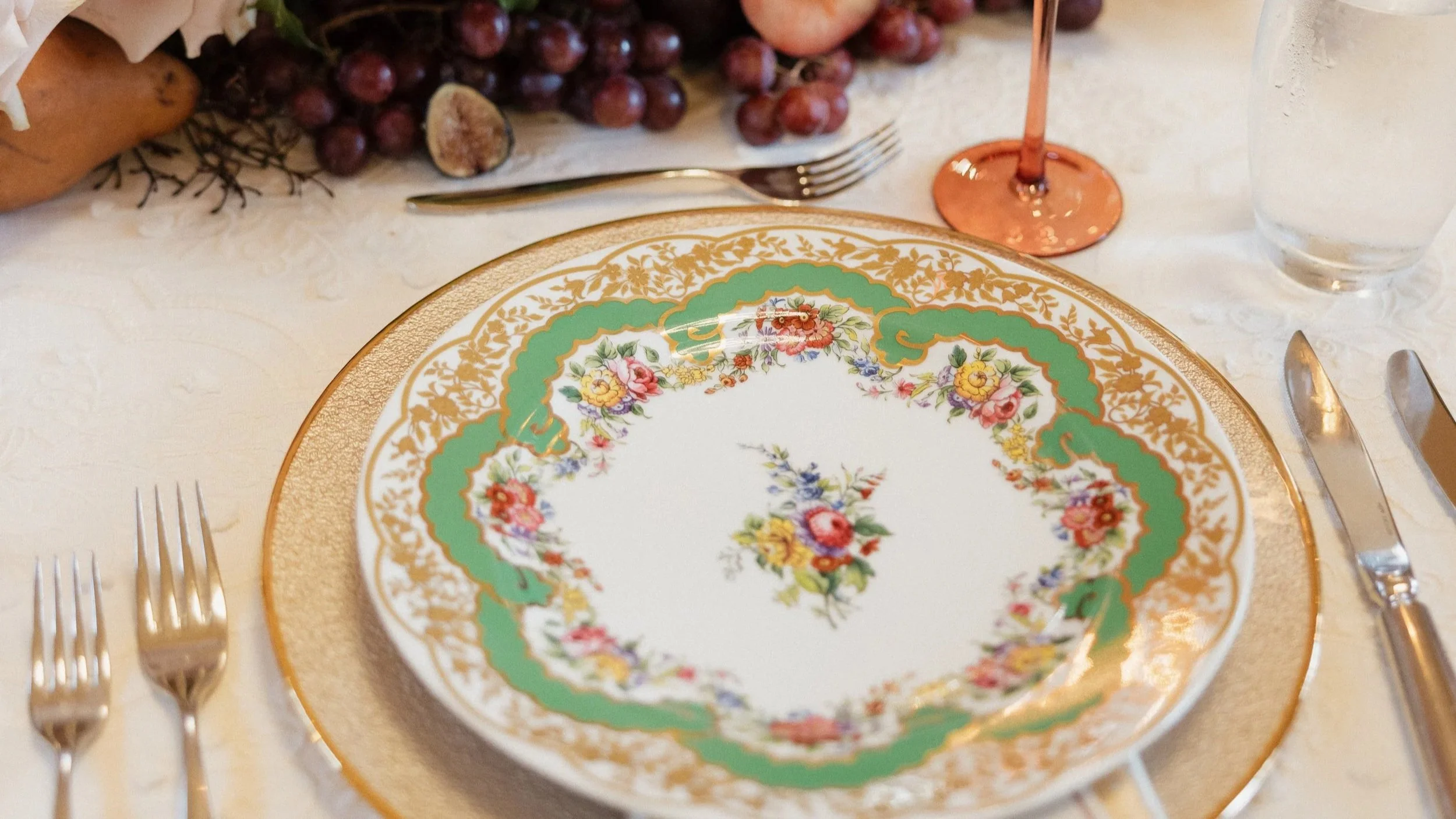 Elegant table setting with a floral patterned plate, gold-rimmed charger, silverware, a glass of water, and a pink drink. Grapes and other fruits are in the background.