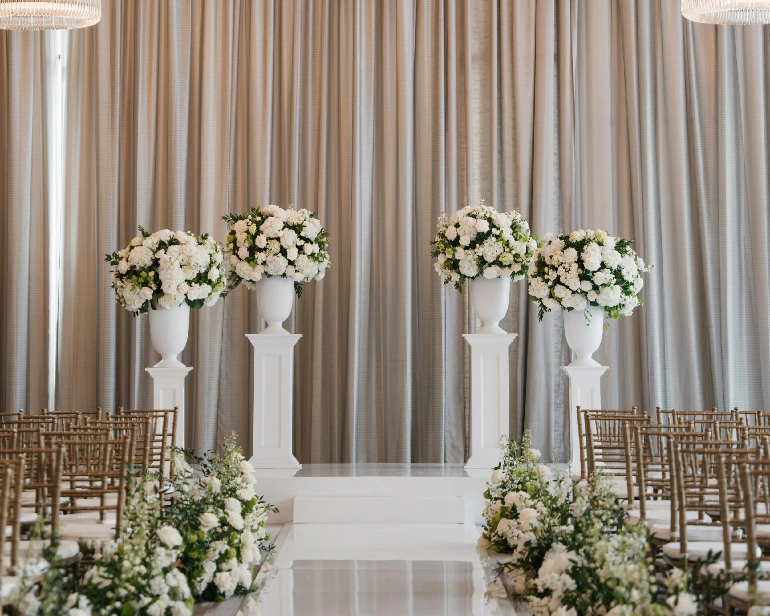Elegant wedding altar decorated with tall white vases holding white floral arrangements, surrounded by white flower arrangements along the aisle, set against a backdrop of beige curtains.