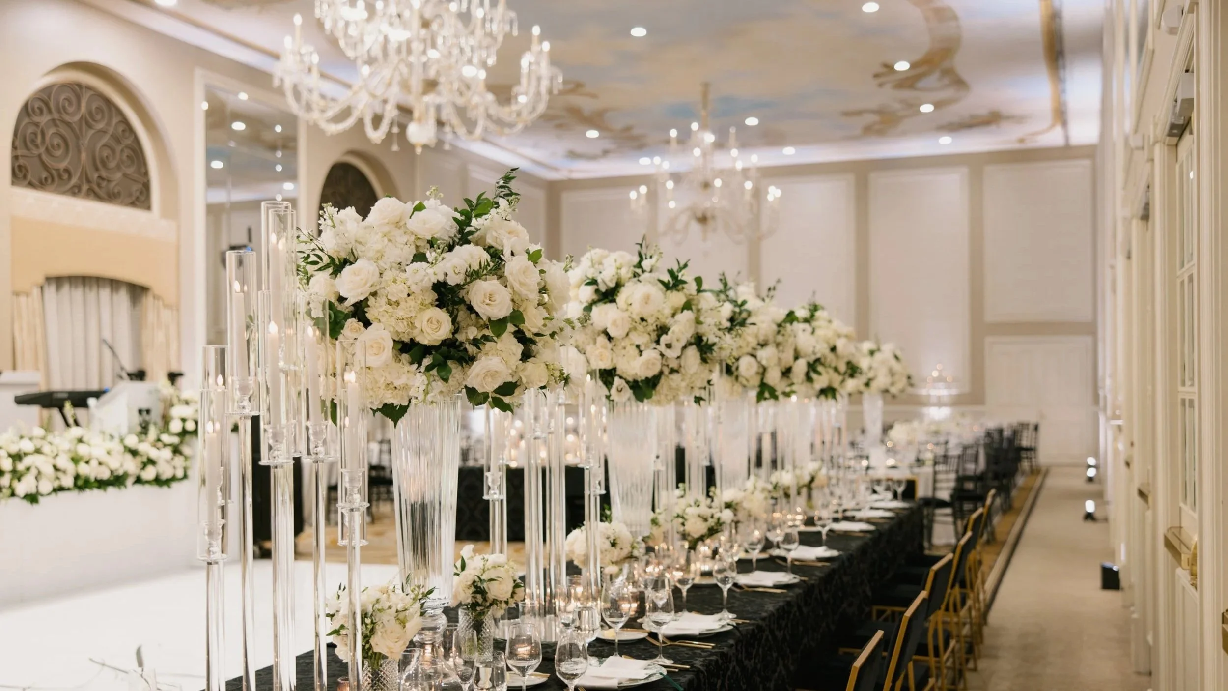 Elegant banquet hall decorated with white floral centerpieces and candles, with chandeliers hanging from a decorated ceiling.