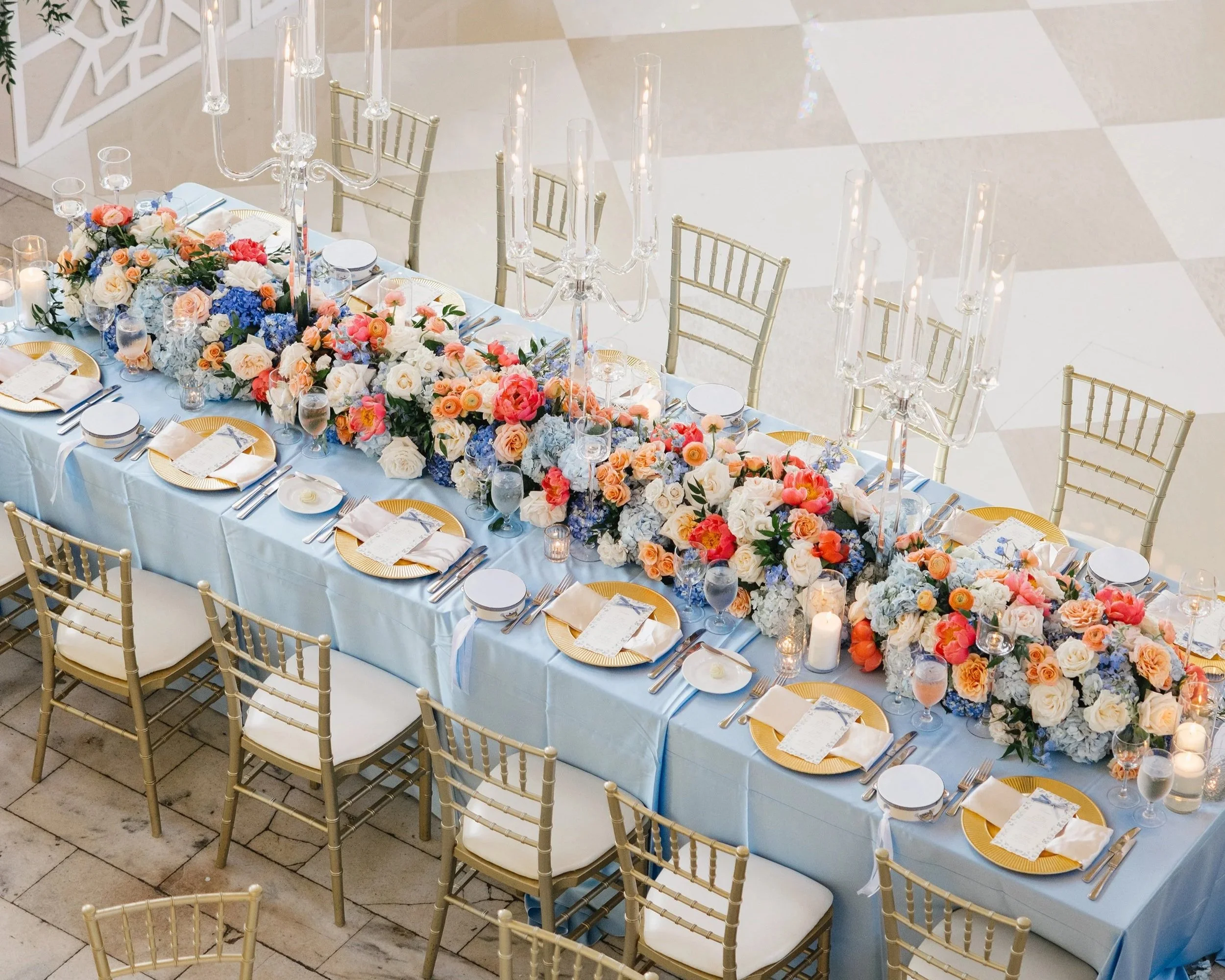 Elegant banquet table decorated with pastel-colored flowers, gold chargers, white napkins, glassware, candles, and tall glass candelabras in a bright, spacious venue.