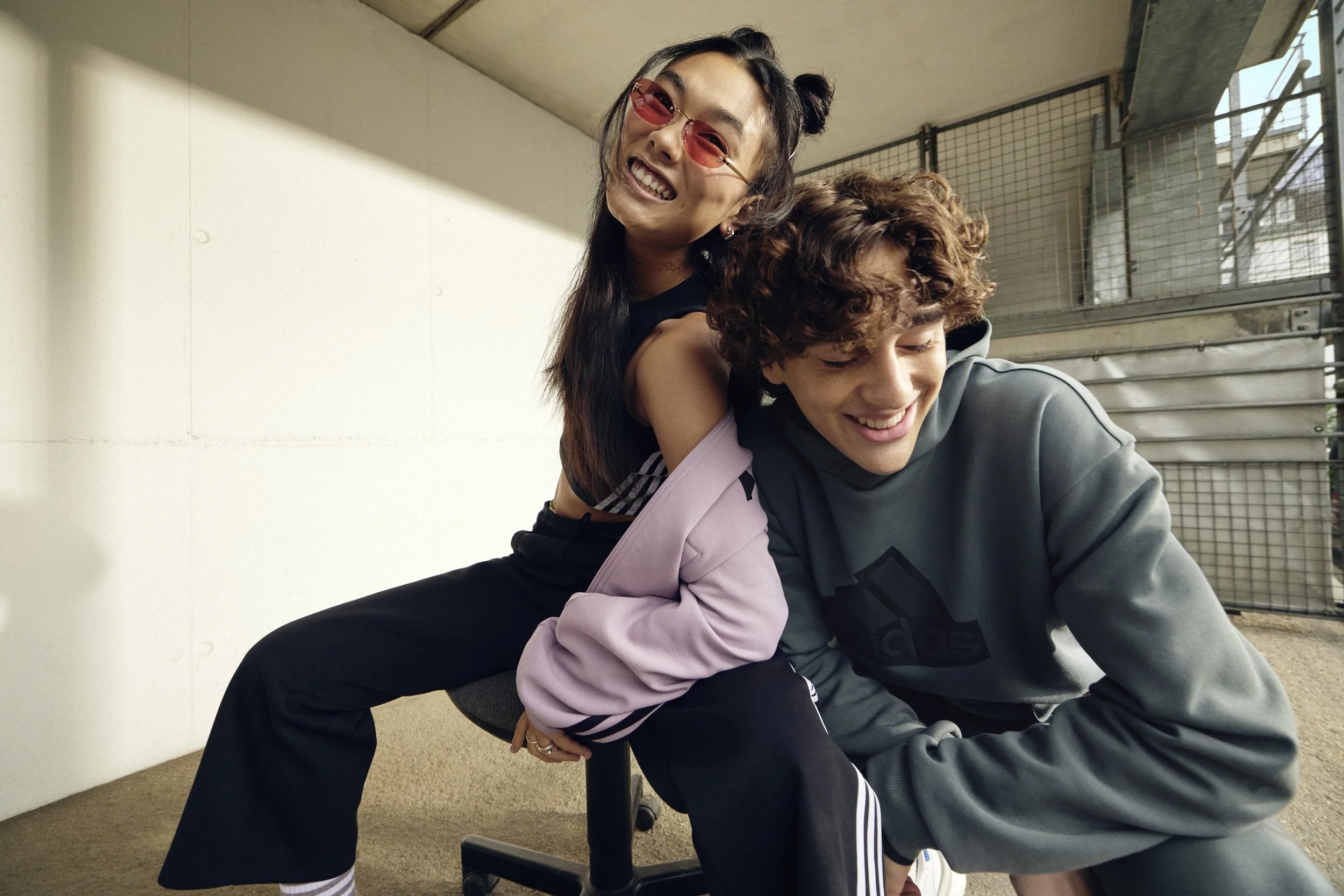 Two young people, a woman with glasses and a man, smiling and sitting closely together in a casual, playful pose indoors with a large window and metal fences in the background.