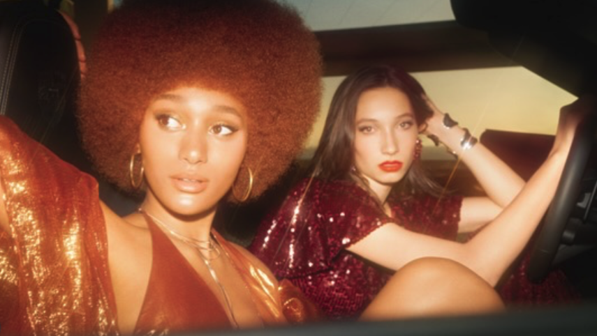 Two young women sitting in a car, dressed glamorously with shiny, sequined outfits, one with an afro hairstyle and the other with long dark hair, both wearing bold makeup and jewelry.