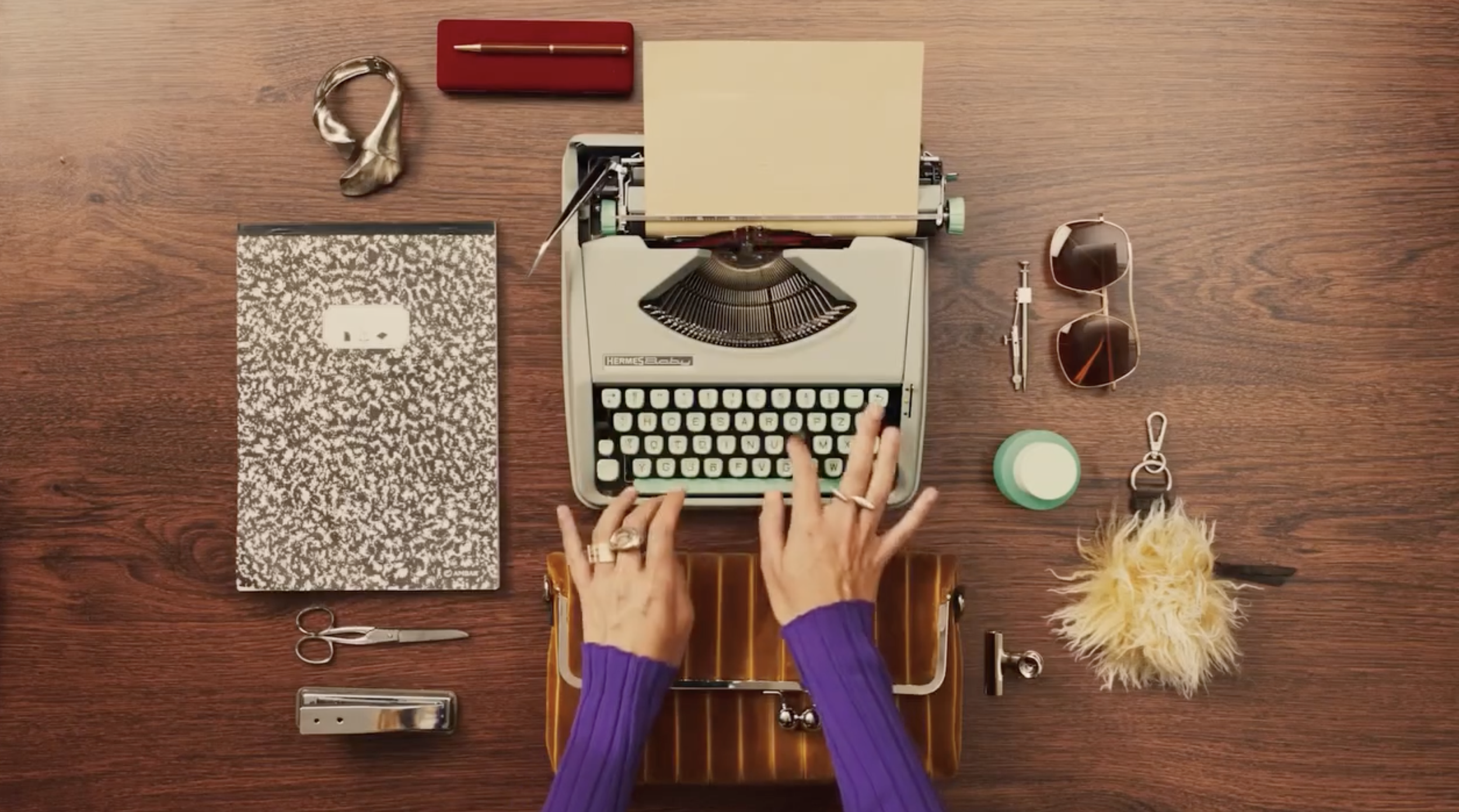 Top-down view of a vintage typewriter on a wooden desk, surrounded by stationery and accessories including scissors, a notebook, sunglasses, a perfume bottle, a fluffy yellow accessory, and a handbag.