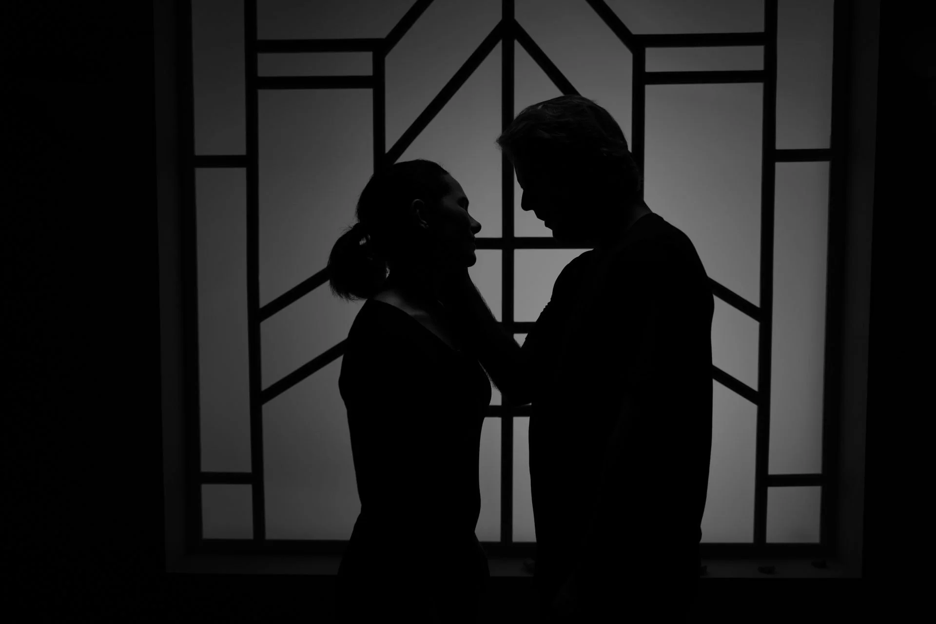 Silhouettes of a man and woman in an intimate pose in front of a decorative window.