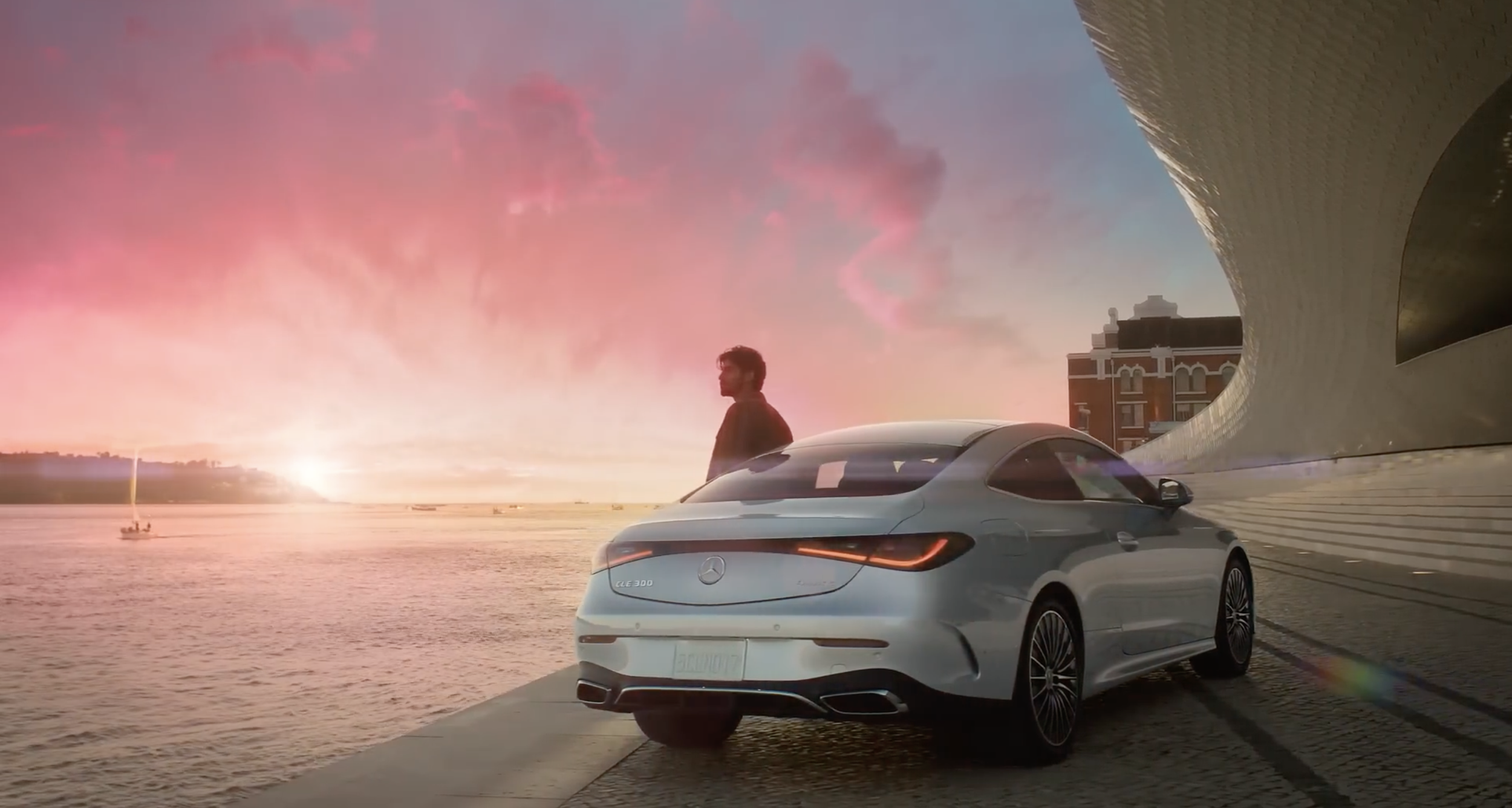 A man standing next to a silver Mercedes-Benz CLE 300 car near a river during sunset with a modern architectural building in the background.
