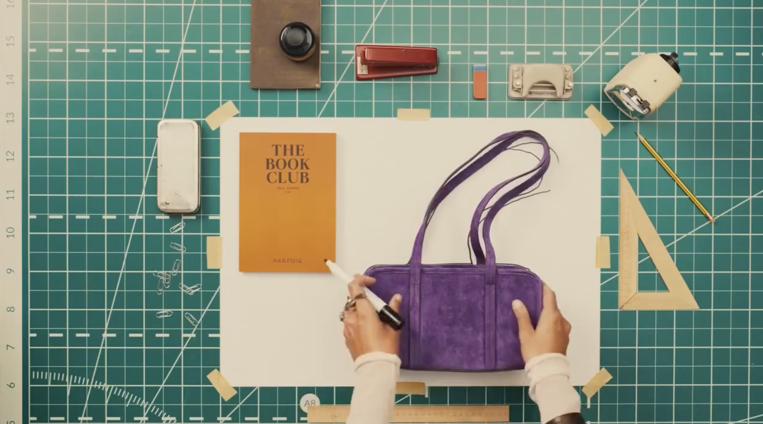 Overhead view of a workspace with a green cutting mat, various tools, a purple bag, a book titled 'The Book Club,' a ruler, a yellow pencil, and a white paper surface.