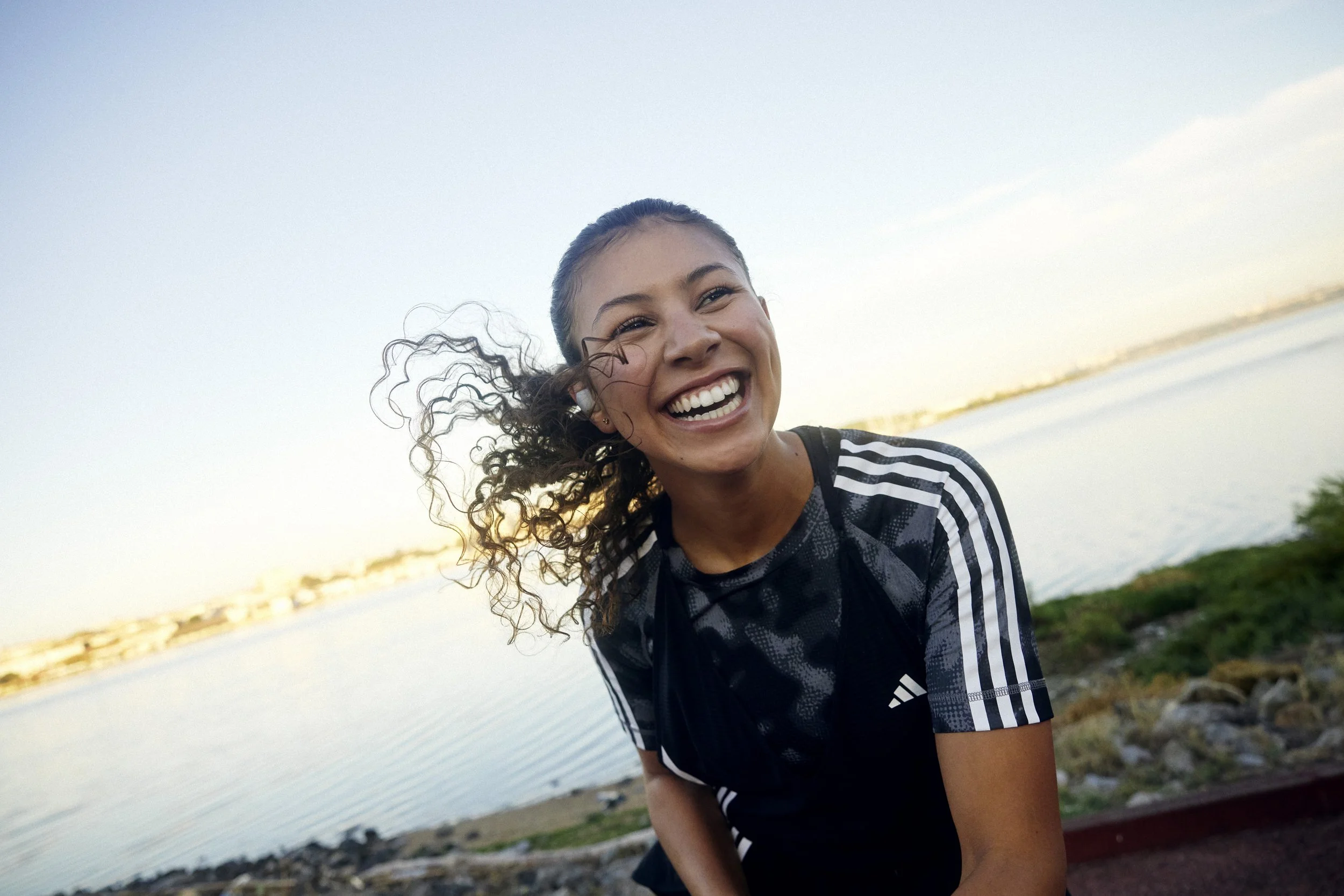 Young woman with curly hair smiling near water with a rocky shoreline during sunset.