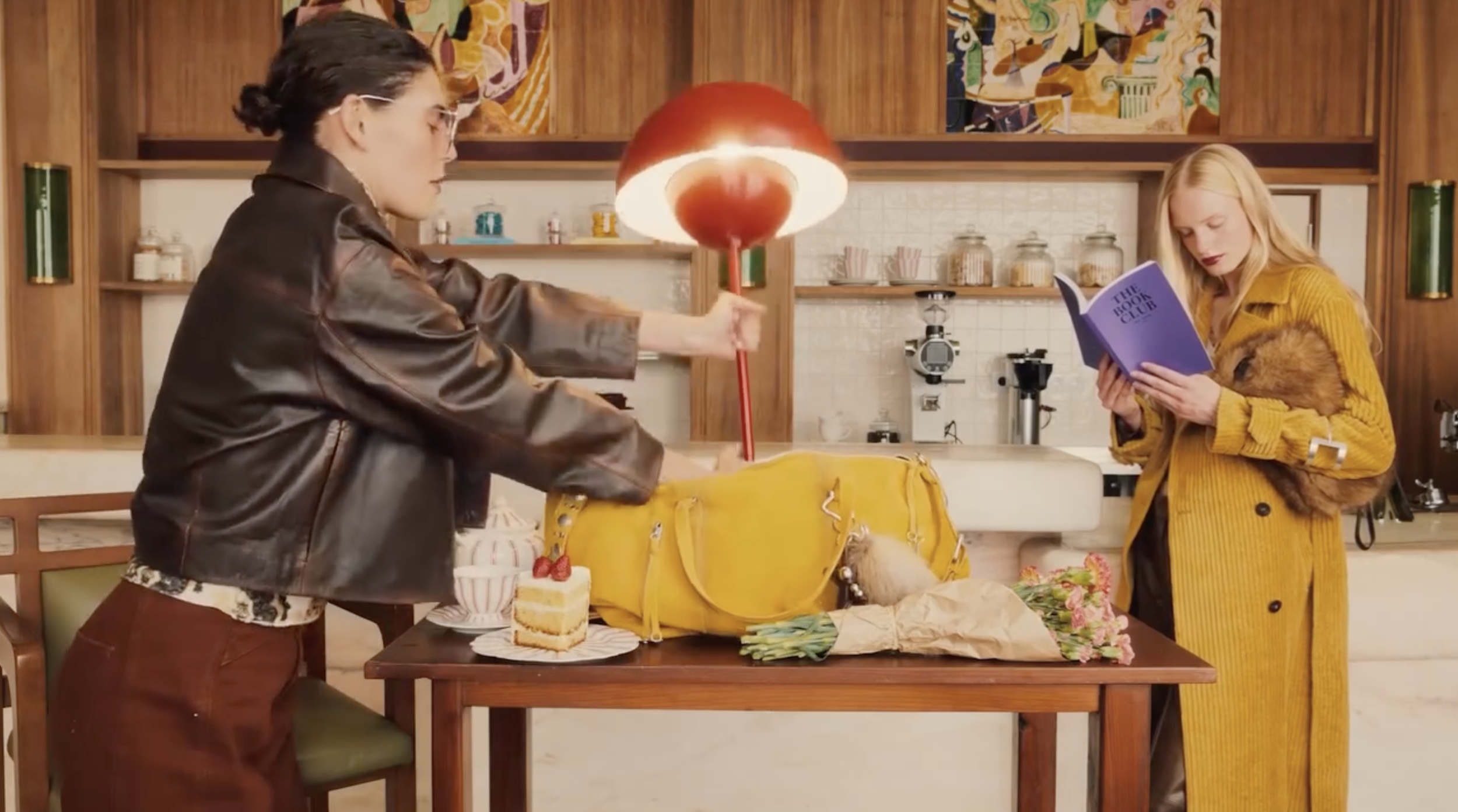 Two women in a cozy kitchen or café, one is reaching into a yellow bag on a table, and the other is reading a purple book titled 'The Book Club' while holding a small dog. There is a birthday cake with strawberries and a bouquet of flowers on the tab