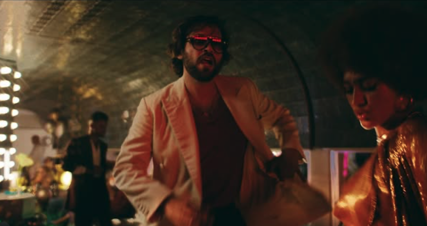 A man with glasses and a beard wearing a beige blazer and red shirt, and a woman with afro hairstyle in a shiny gold jacket, are in a dimly lit nightclub or party setting with other people in the background.