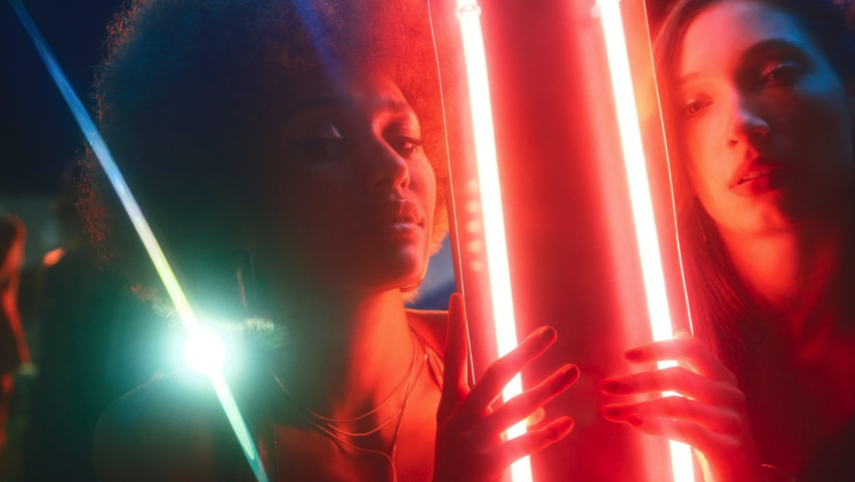 Two women in a dark setting illuminated by colorful neon lights, one holding a glowing cylindrical object.