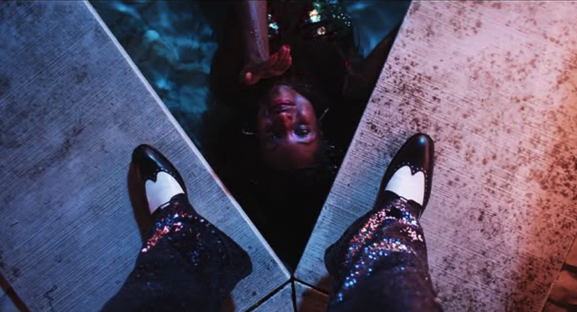 Person lying on the floor with their head between two mirror edges, seen from the perspective of someone standing and looking down at their shoes and legs.