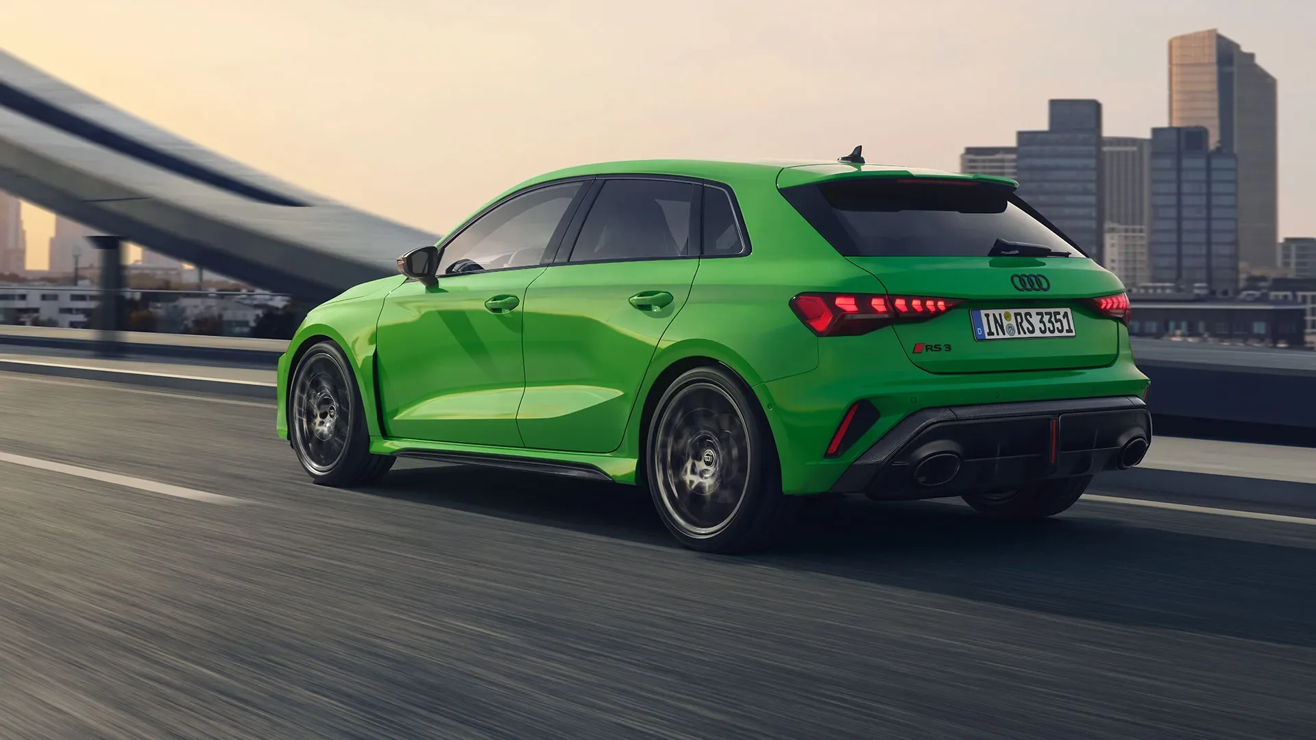 A bright green Audi RS 3 hatchback driving on a city highway with buildings in the background.