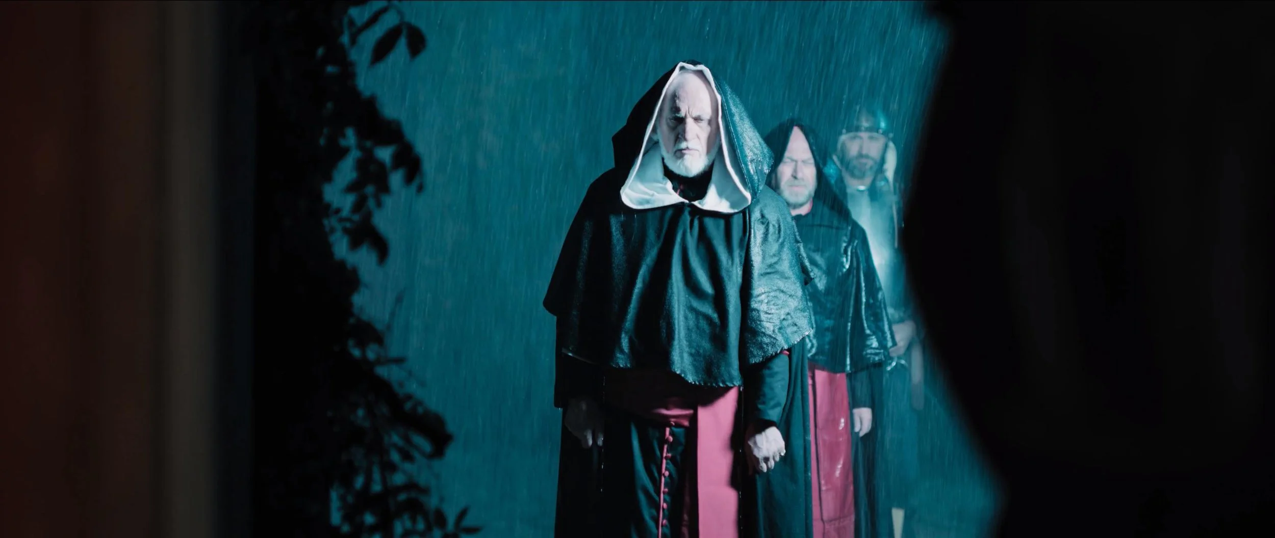 Three men standing in the rain at night, seen through a partially open door or window. The man in front wears a black hooded cloak with a white lining and has a serious expression. The other two men stand behind him, also in dark clothing, with serio