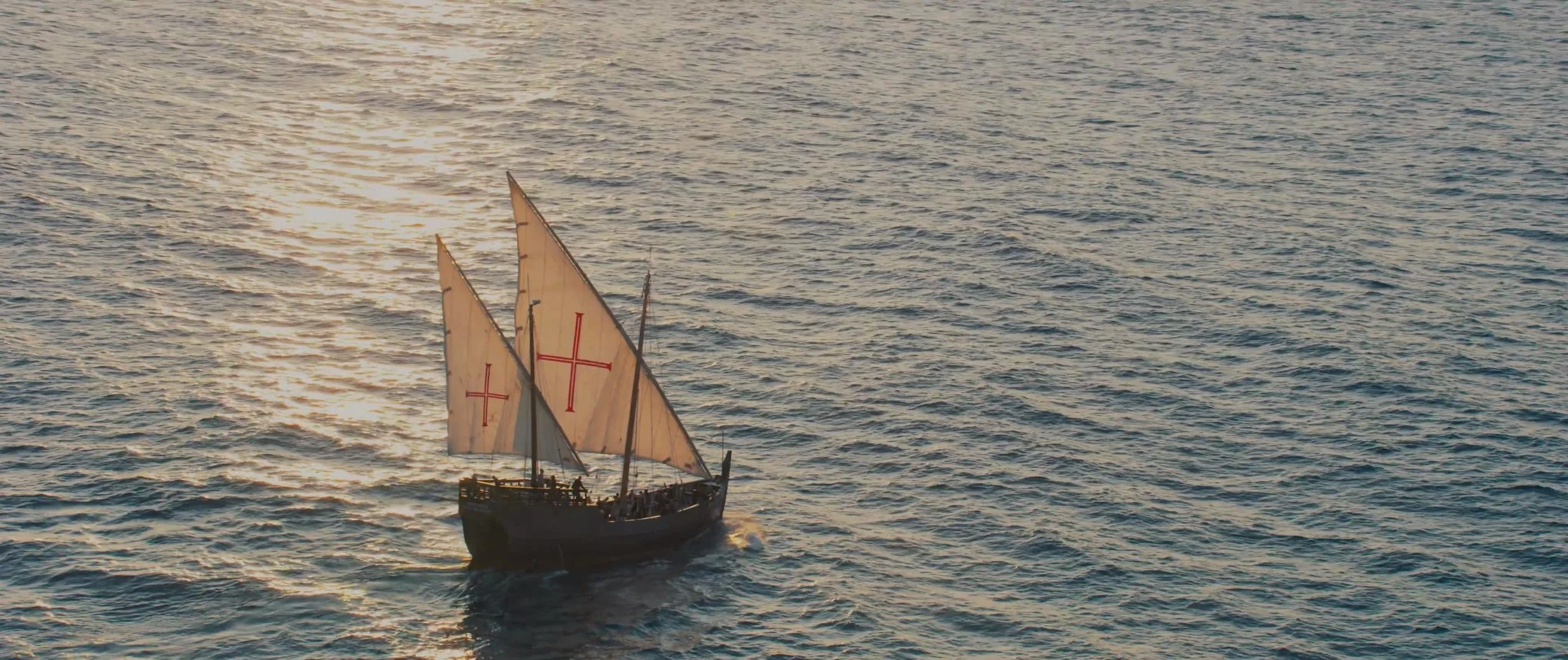 A large sailing ship with three sails featuring red crosses, cruising on the ocean during sunset.