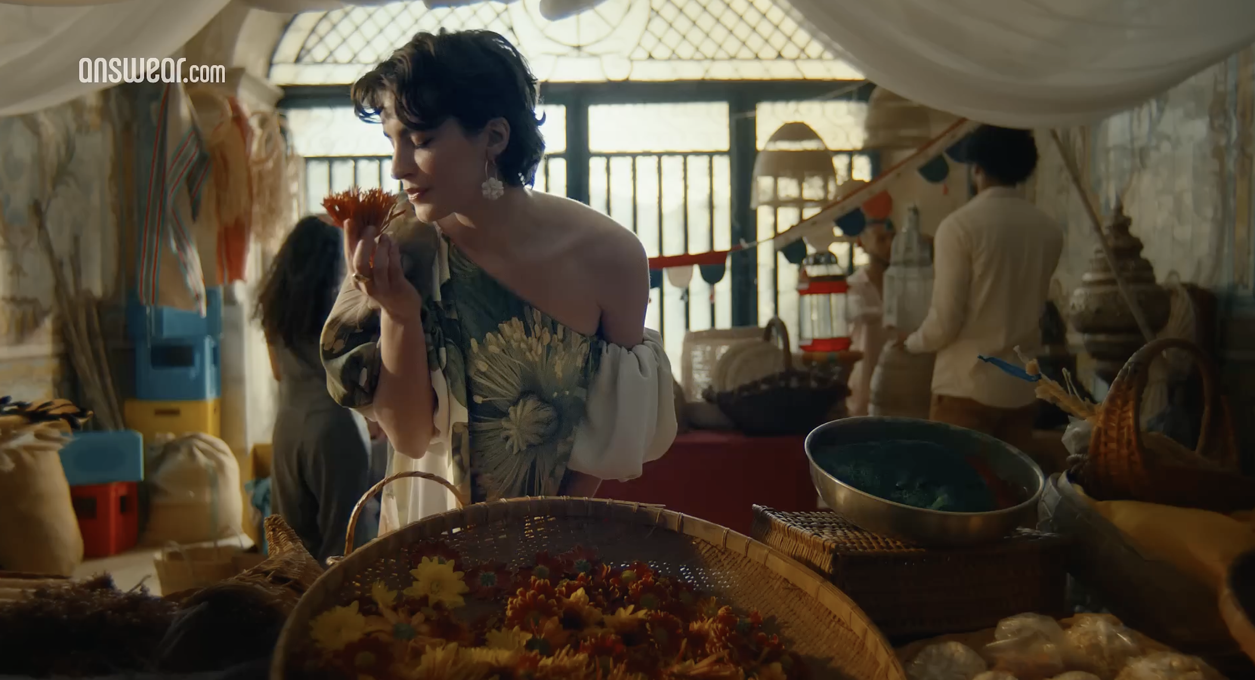A woman with short dark hair, wearing a floral off-shoulder dress and large earrings, smells a flower in a marketplace with baskets of flowers and other goods, with people in the background.