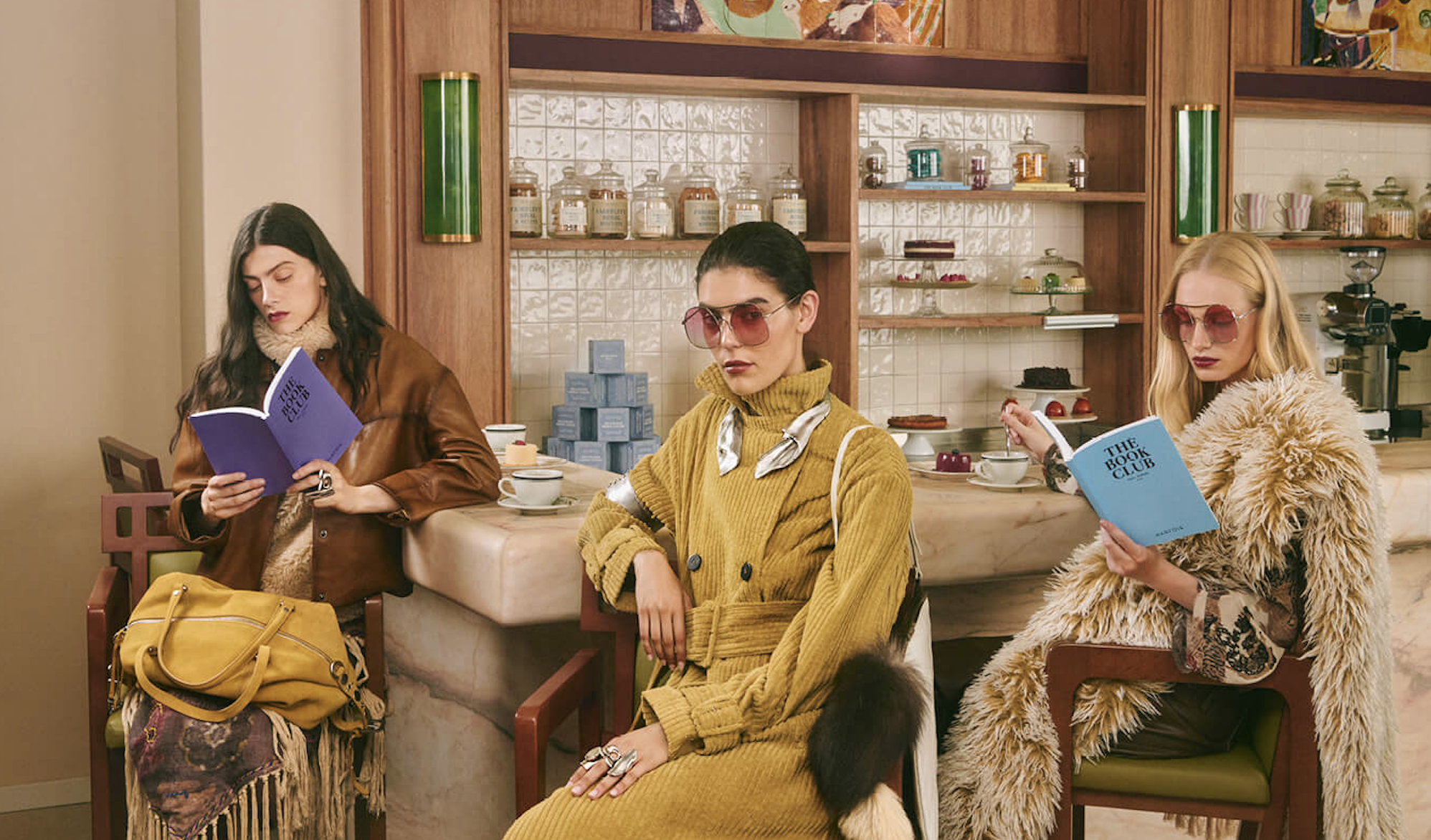 Three women sitting in a vintage-style coffee shop. The woman on the left has long dark hair, is wearing a brown leather jacket, and is reading a purple book titled 'The Book Club.' The woman in the middle has short dark hair, wears a yellow sweater 