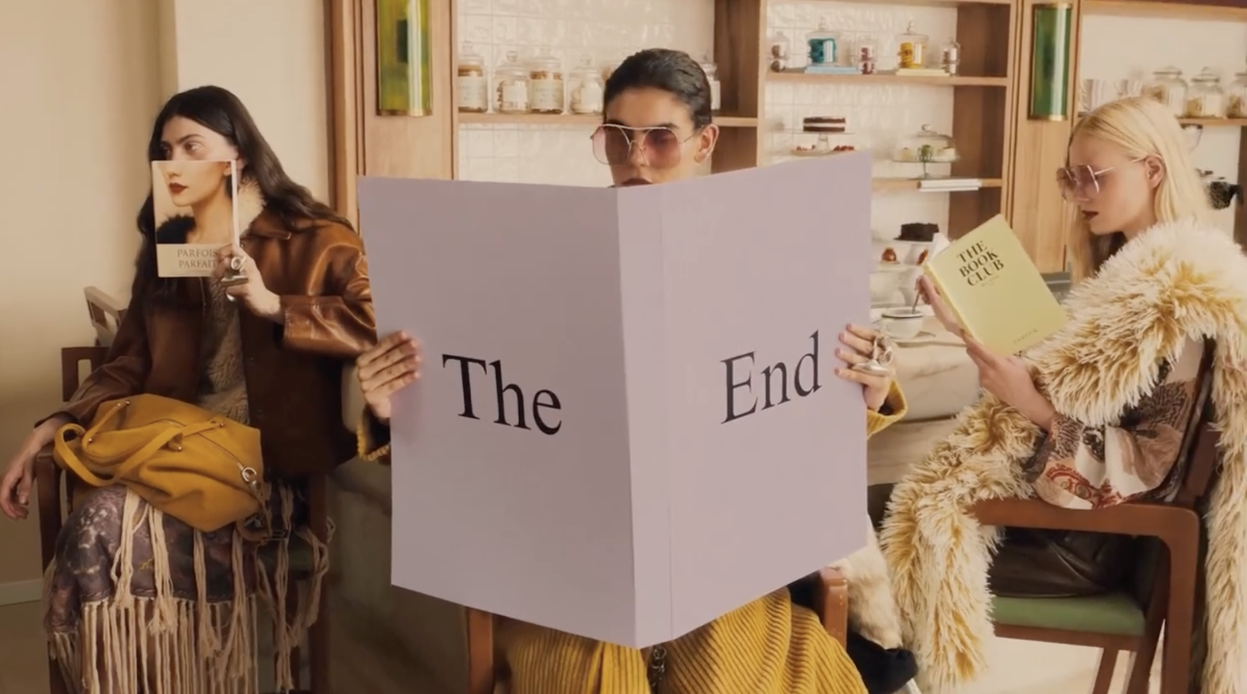 Three women sitting at a table in a cafe, holding books and looking in different directions. The woman in the middle is reading a large sign that says "The End".