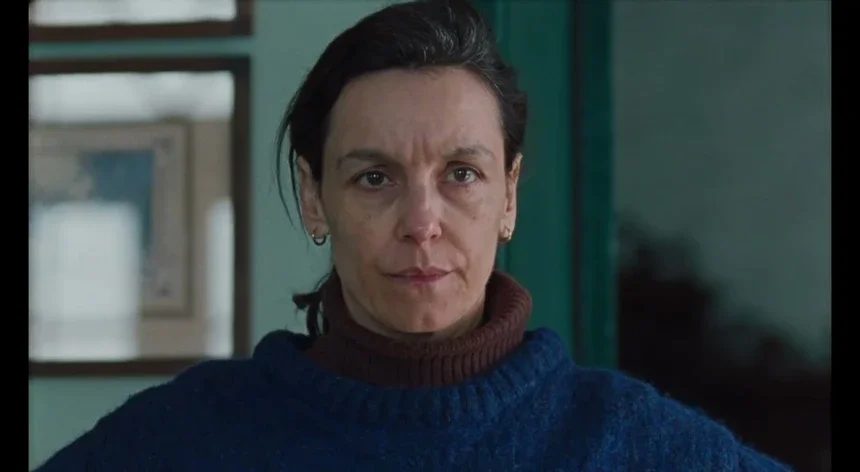 Close-up of a woman with short dark hair wearing a dark blue sweater and a brown turtleneck, looking seriously at the camera.