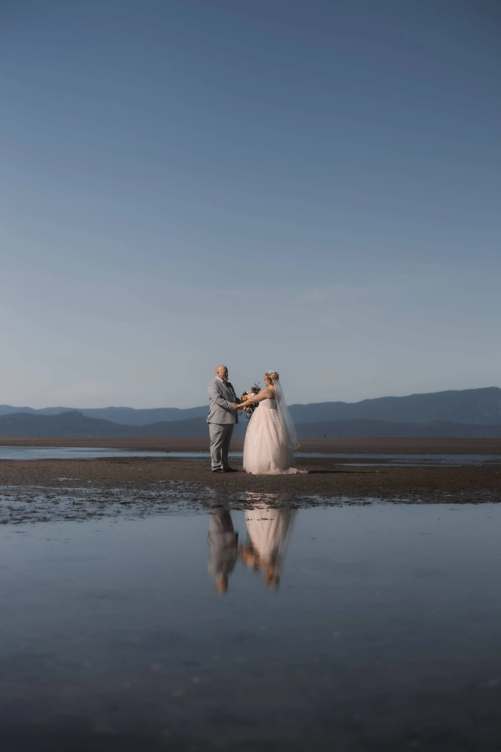 Newlyweds reflected in shallow water during beach portraits in Parksville, BC