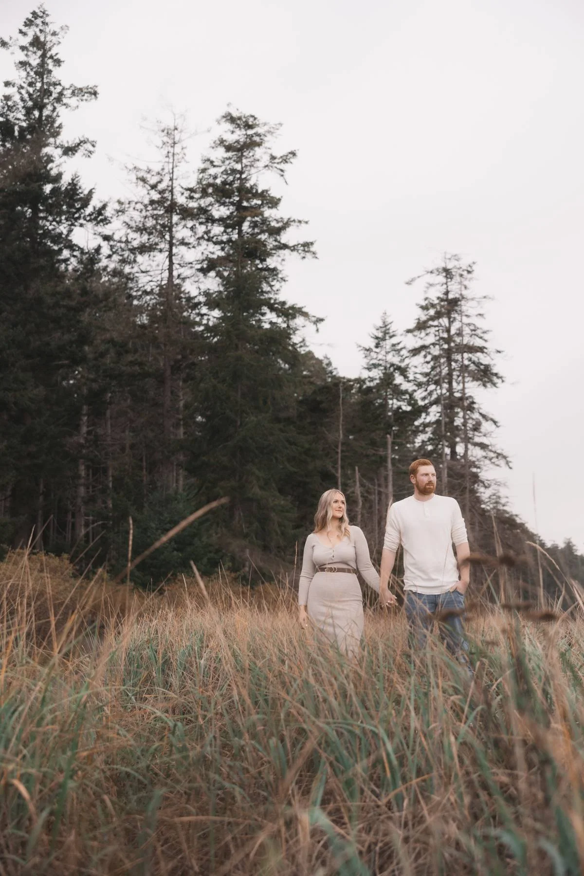 Couple walking through coastal grasses near the forest during an engagement session in Parksville BC