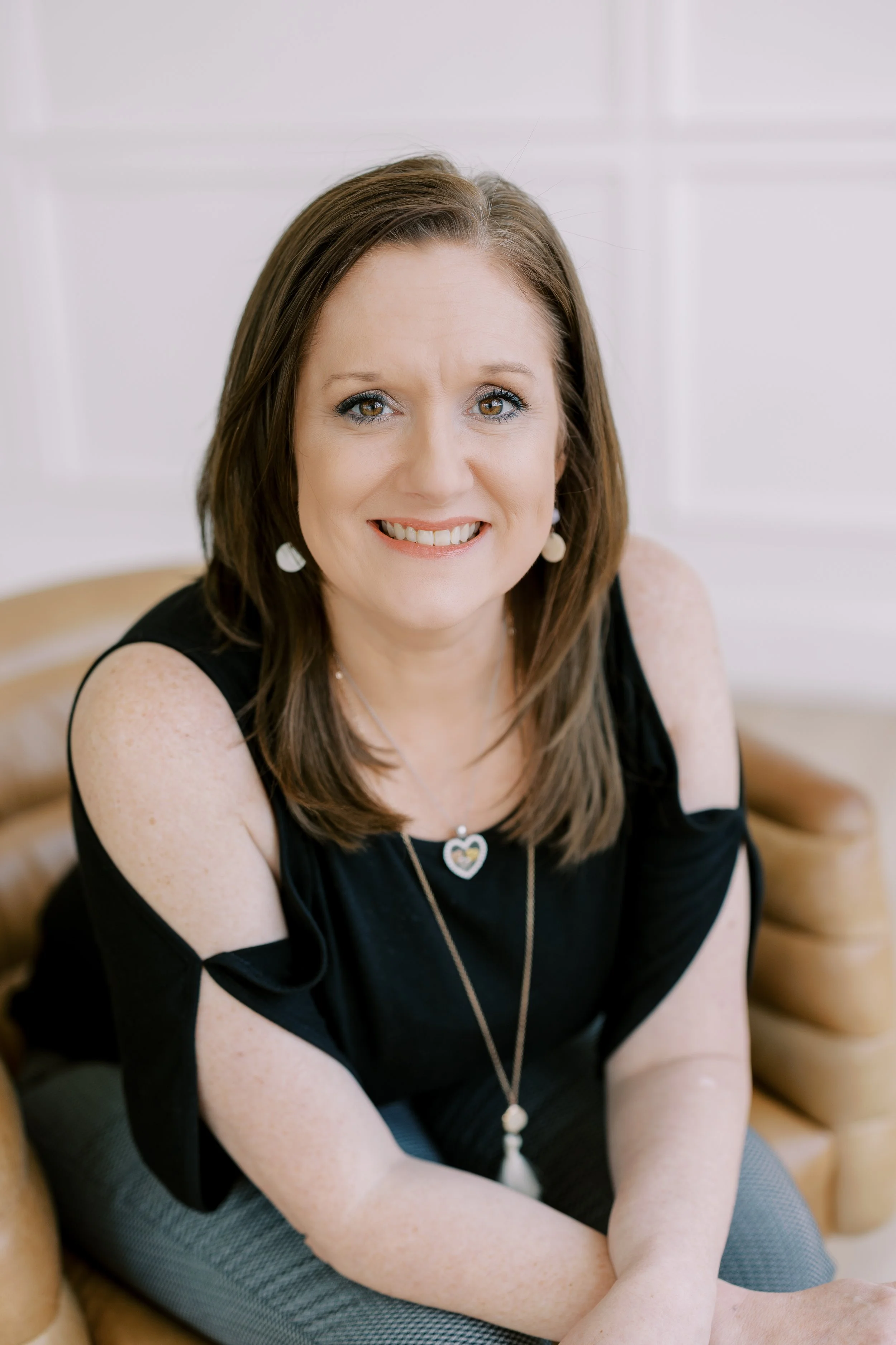 A woman with shoulder-length brown hair, wearing a black top with cutout shoulders and jewelry, sitting on a tan leather couch in a white room, smiling at the camera.