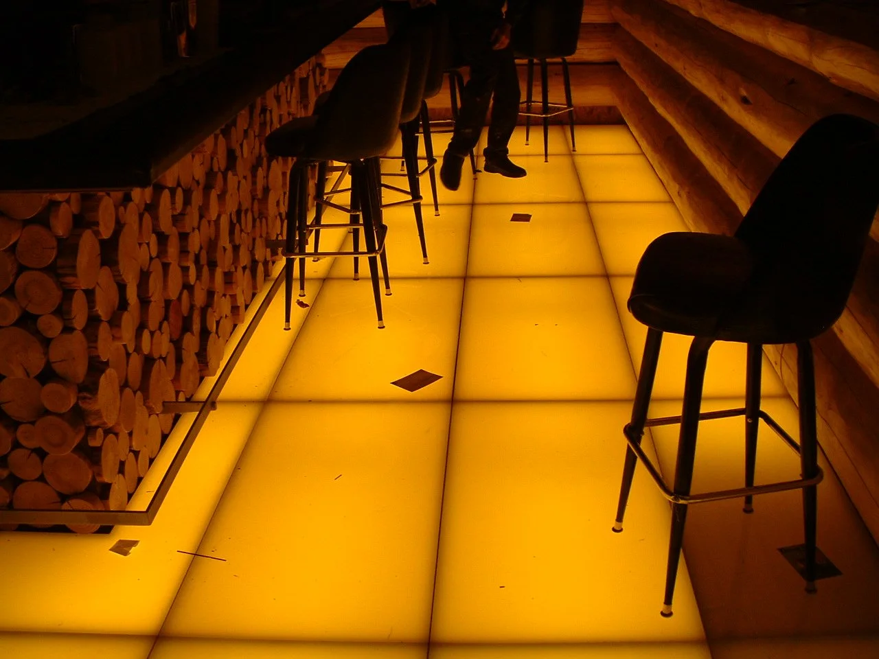 Interior of a bar with a glowing yellow tile floor, wooden log walls, a row of bar stools, and a person sitting at the bar.