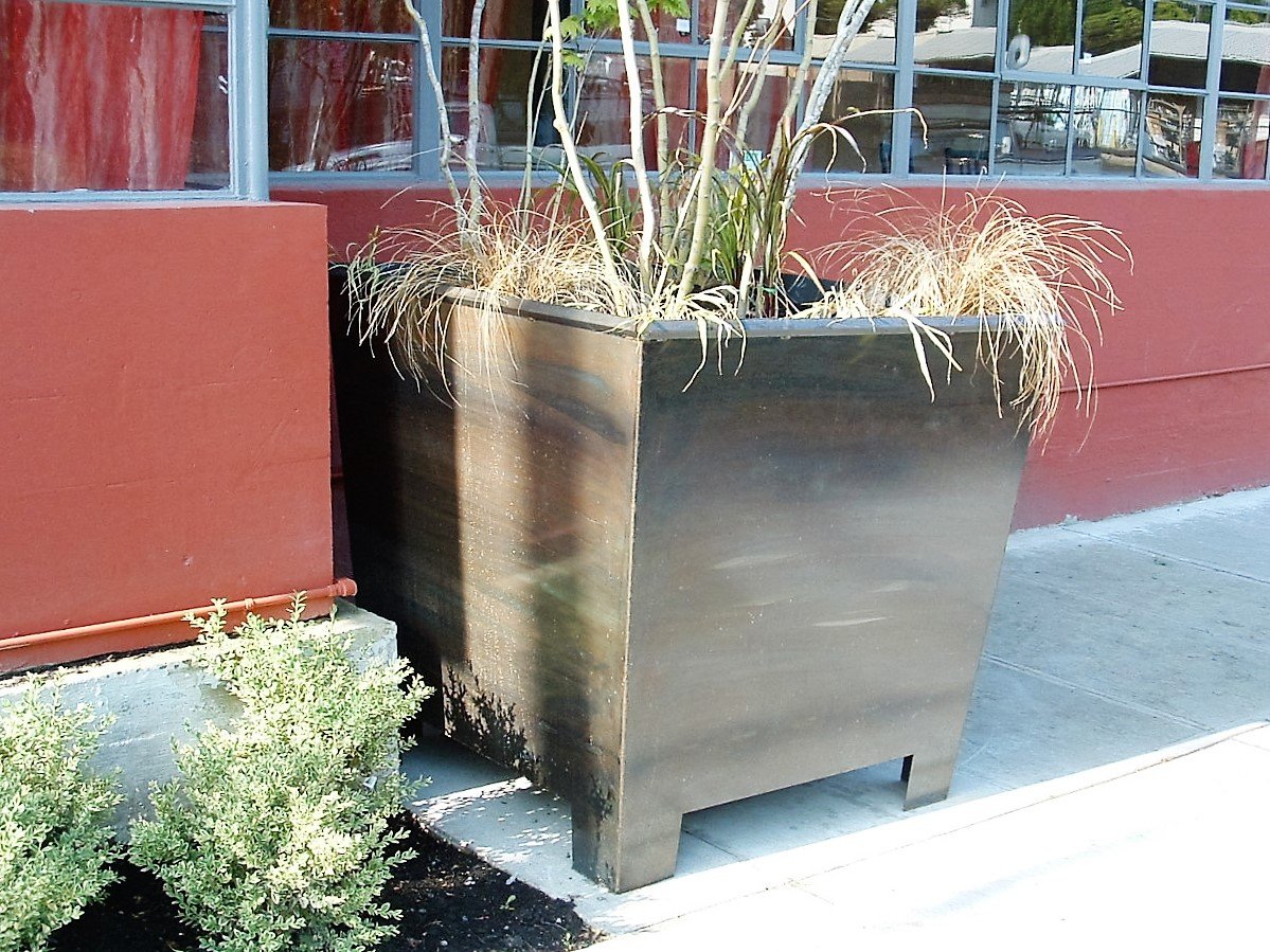 Large black metal planter with plants, positioned outside a building with large windows and a red exterior wall.
