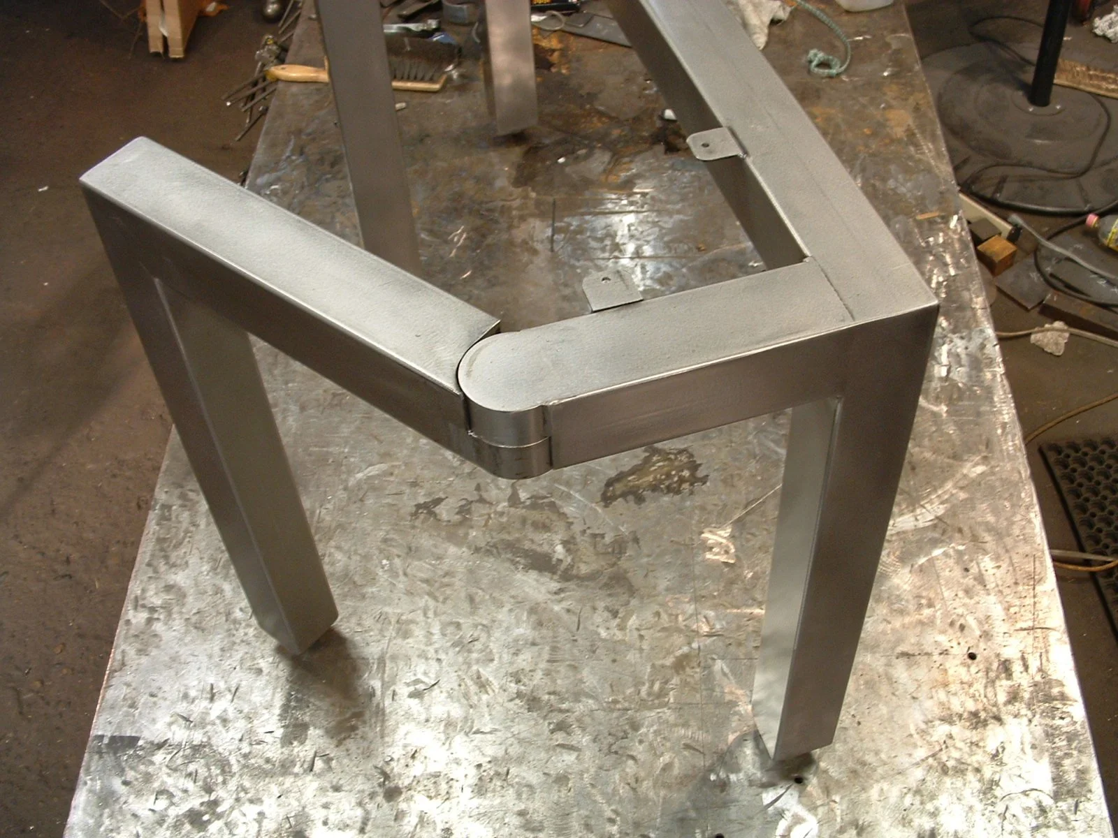 A metallic table with an unfinished metal frame on it, in a workshop setting with tools and equipment in the background.