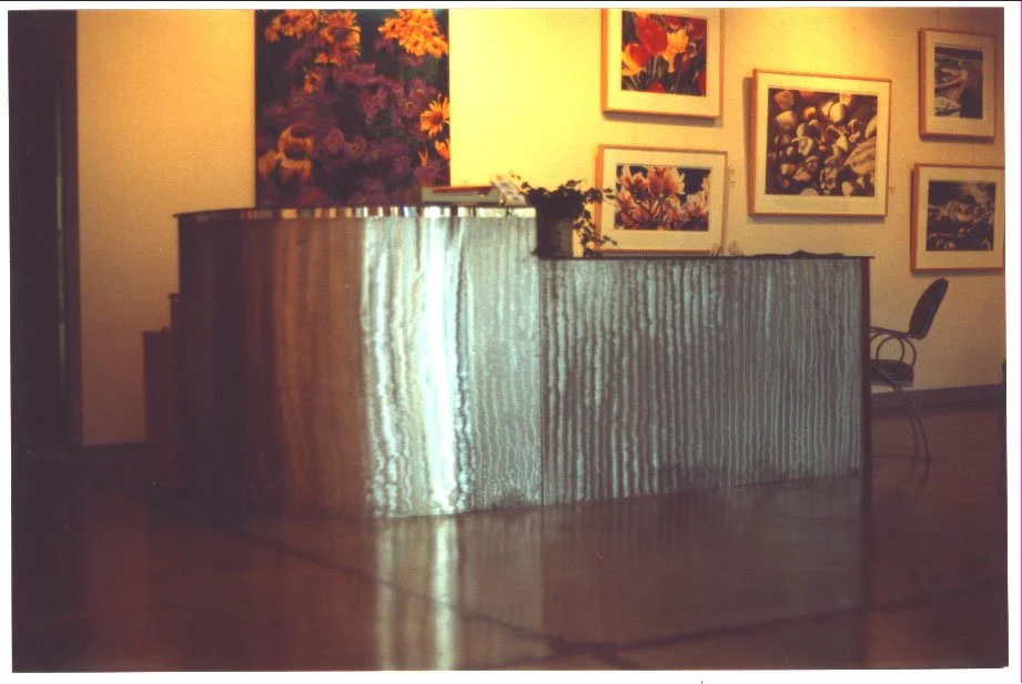 An indoor art gallery or showroom featuring abstract and floral paintings on the wall and a metallic reception desk with a potted plant and some books on top. There is a black chair nearby.