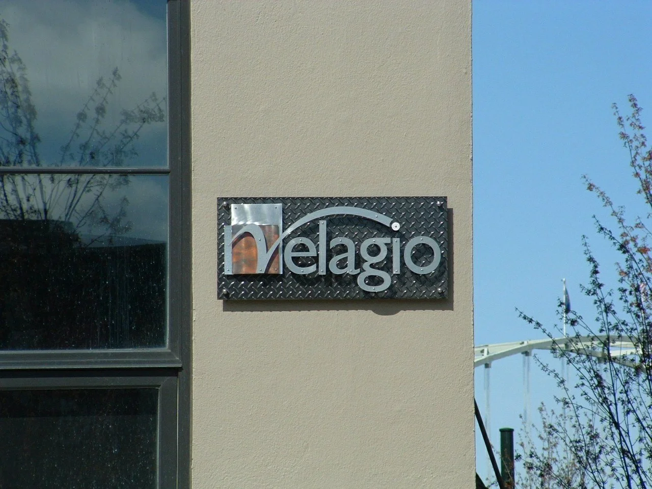 Metal and glass sign on beige wall displaying the word 'Velagio' with a stylized 'm' and a curved line above it, next to a window reflecting trees and a blue sky.