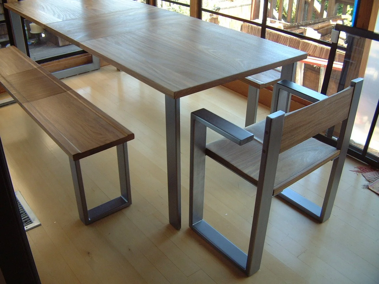 Wooden dining table with metal legs, a matching bench, and a chair with wooden armrests in a room with hardwood flooring and large sliding glass doors to a patio.