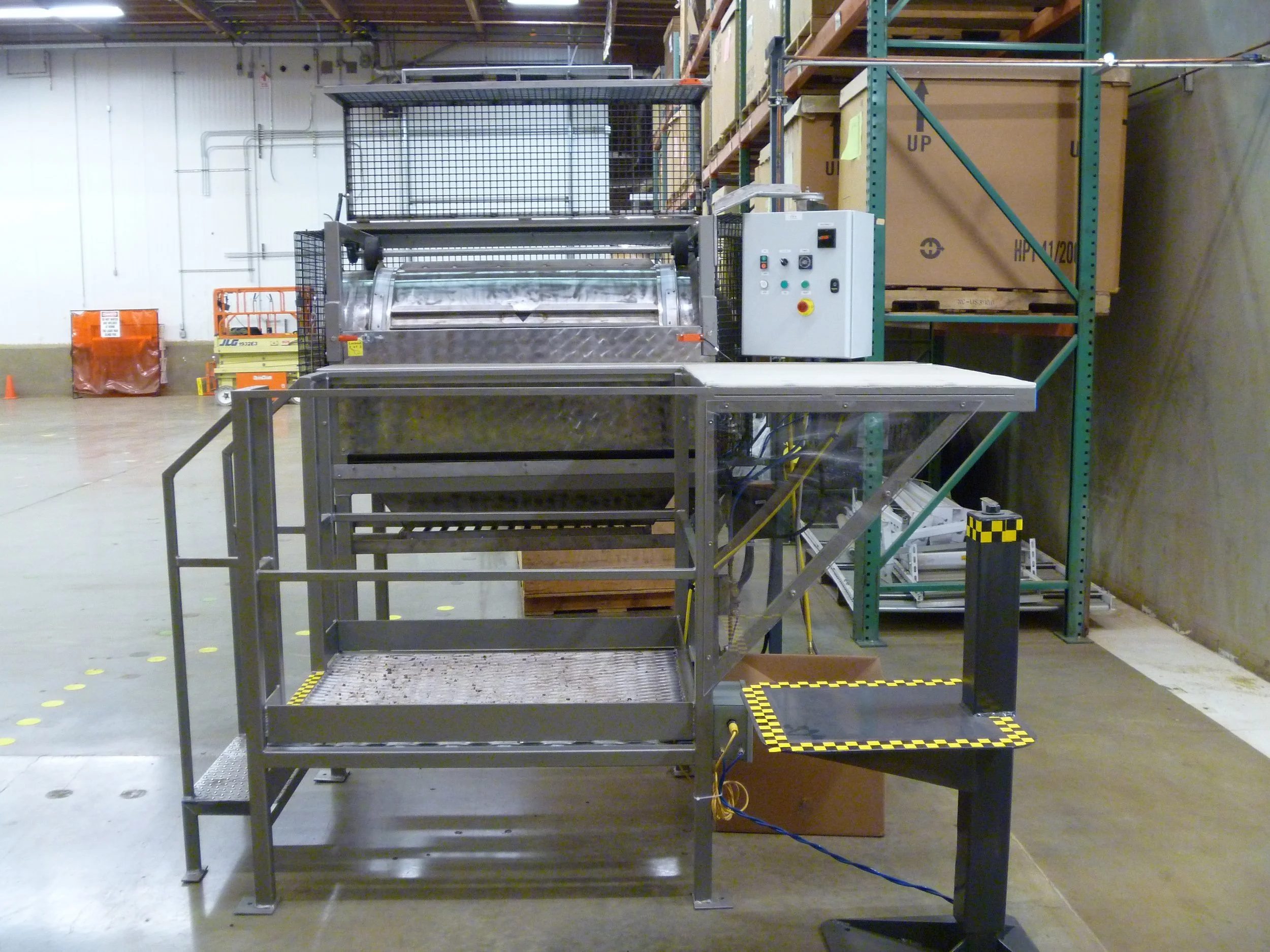 Industrial stainless steel machine inside a warehouse, with control panel, shelving, and cardboard boxes in the background.