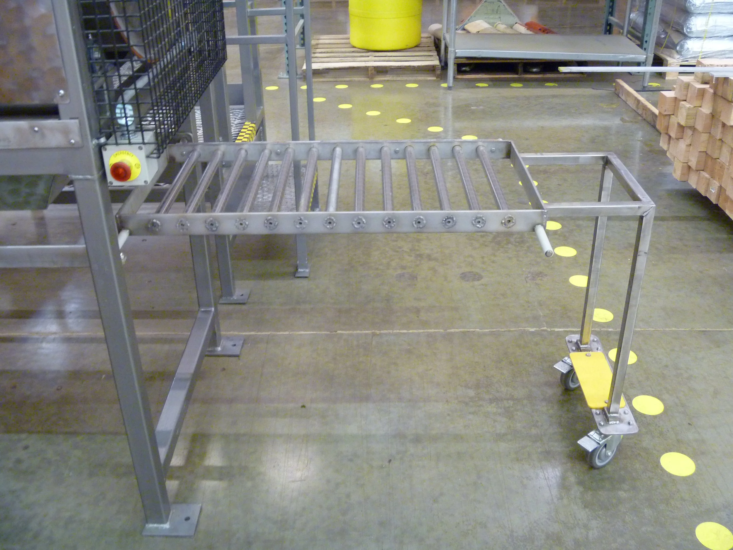 A metal roller conveyor table with yellow caster wheels in a warehouse or manufacturing setting, surrounded by storage racks and pallets.
