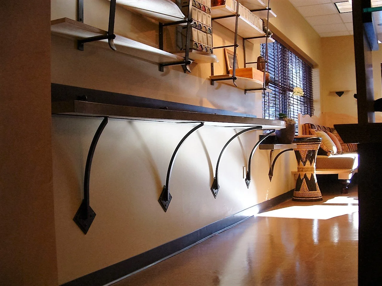 Empty wall-mounted shelf with brackets in a cozy living room, featuring a sofa, decorative pillows, a woven side table, potted plants, and a large window with blinds.