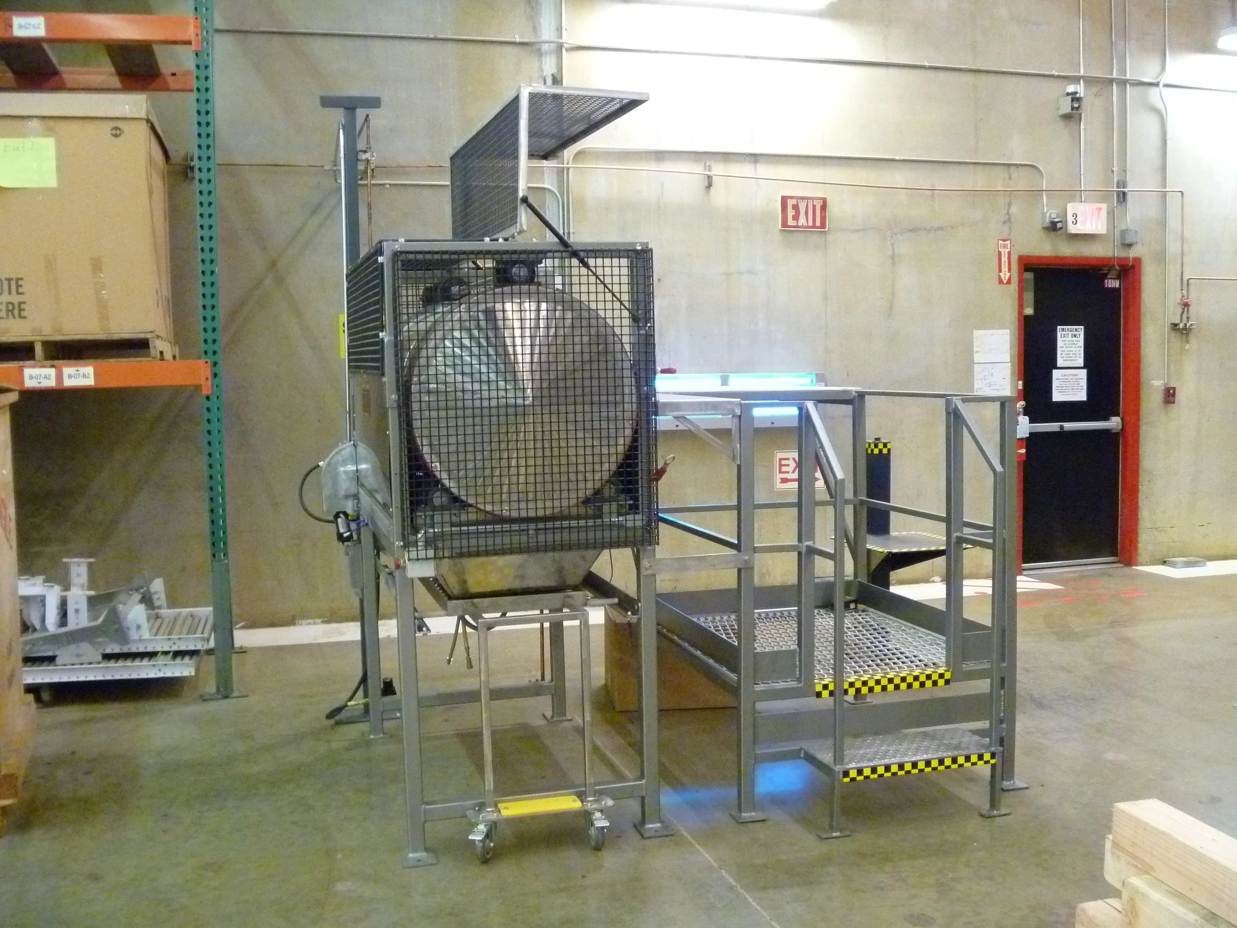 Industrial warehouse with metal equipment, a large cylindrical device enclosed in a protective cage, and an emergency exit door in the background.