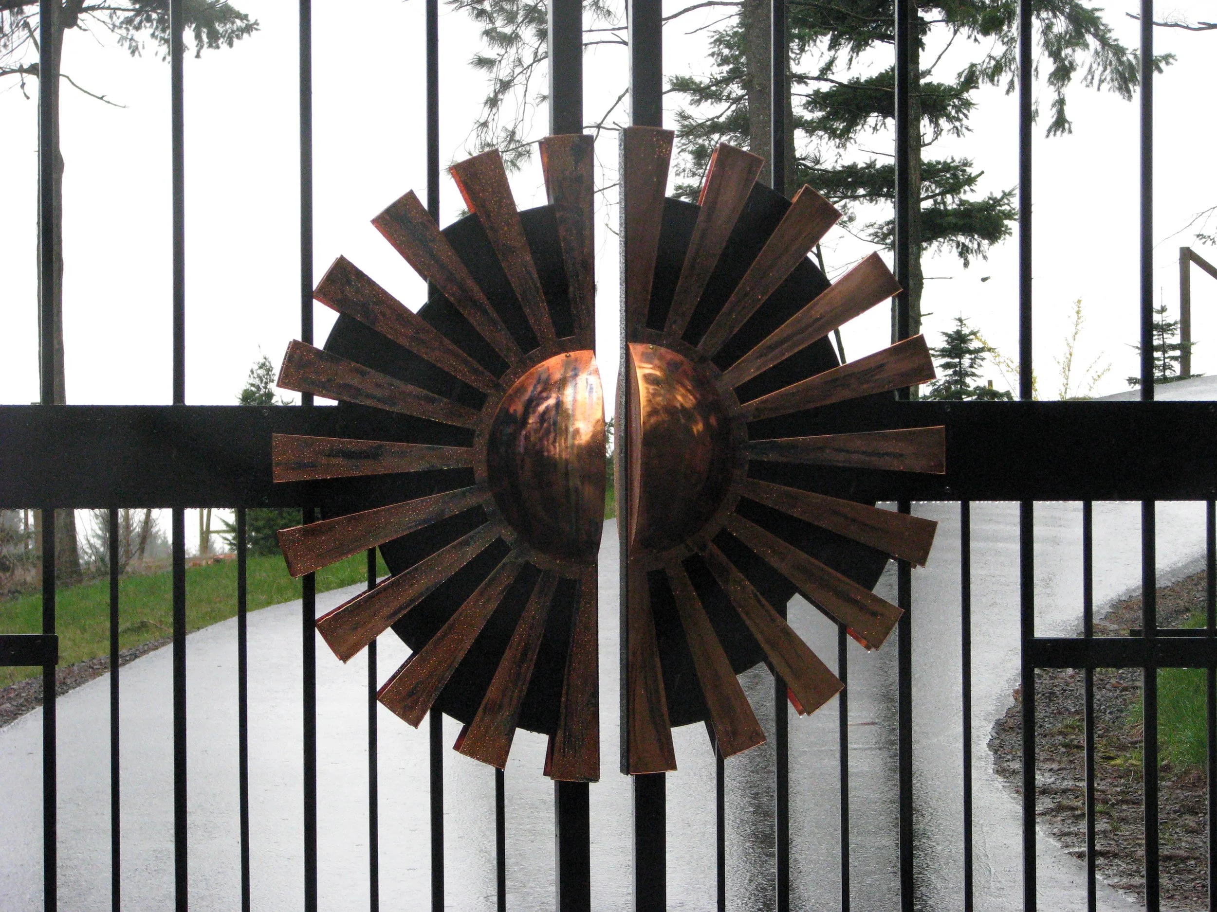 Decorative metal sunburst sculpture mounted on a black metal gate overlooking a wet paved driveway with trees in the background.