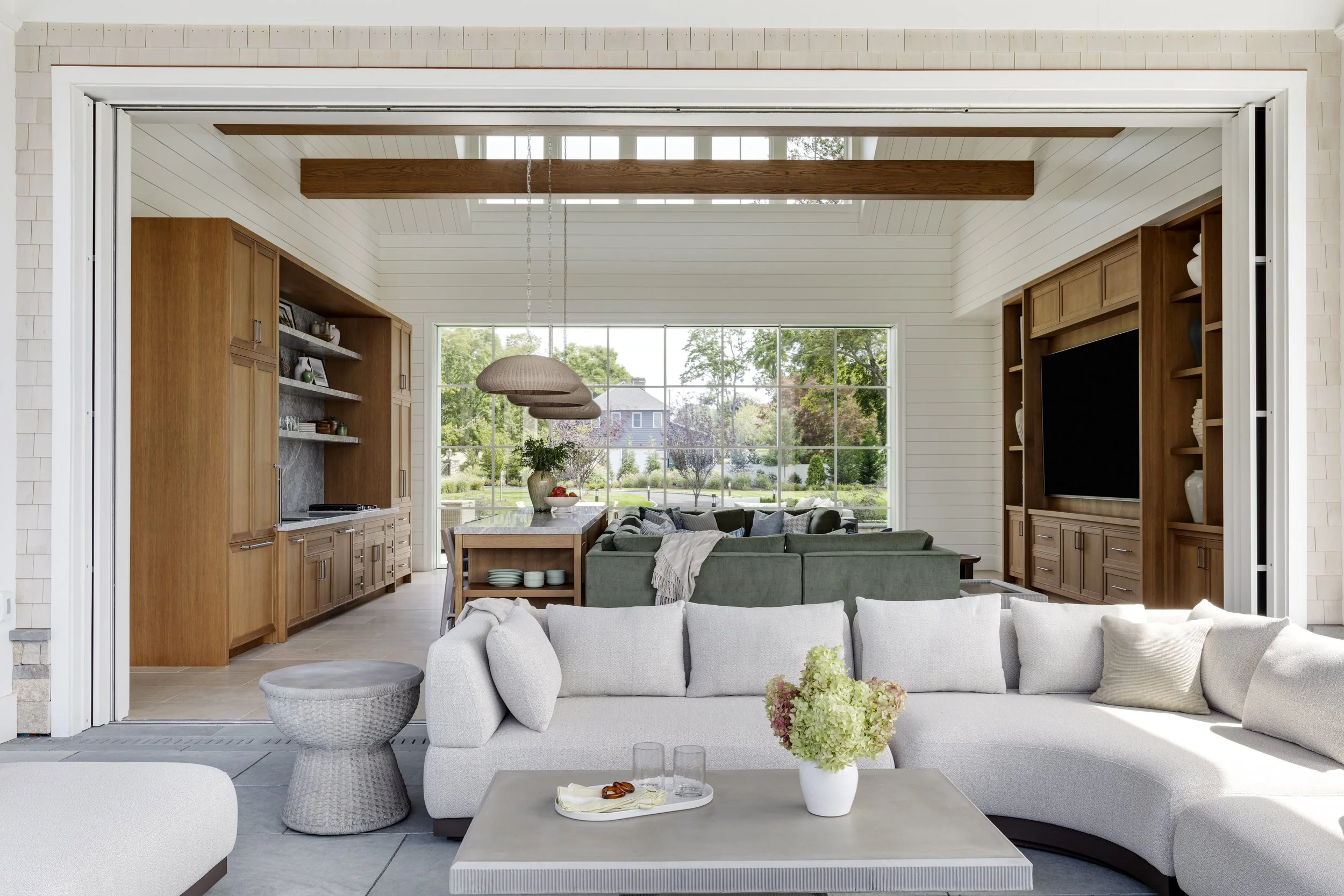 Luxury new construction pool house in Warwick, Rhode Island, featuring an open-air lounge with curved sectional seating, custom wood cabinetry, exposed ceiling beams, and seamless indoor-outdoor living.