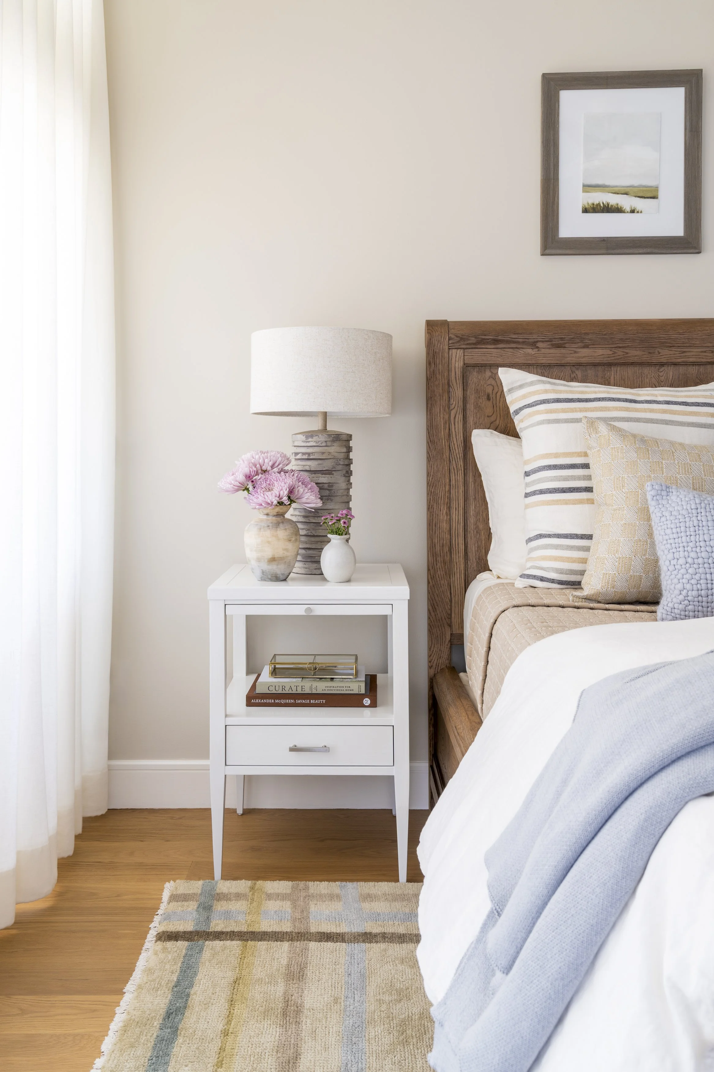 Coastal bedroom bedside detail with woven wood bed, white nightstand and layered blue and neutral textiles in Narragansett, RI.