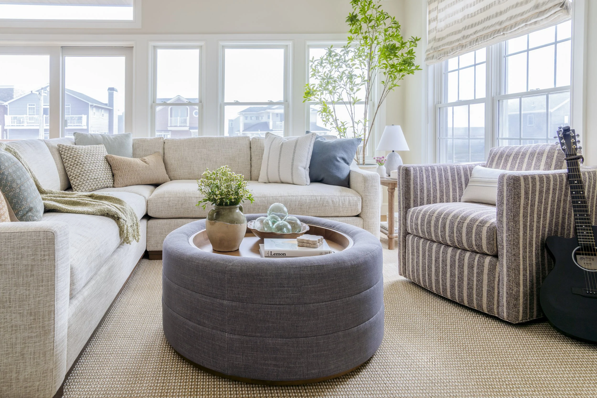 Light-filled family room with layered neutral furnishings and a round textured ottoman in a Narragansett beach house.